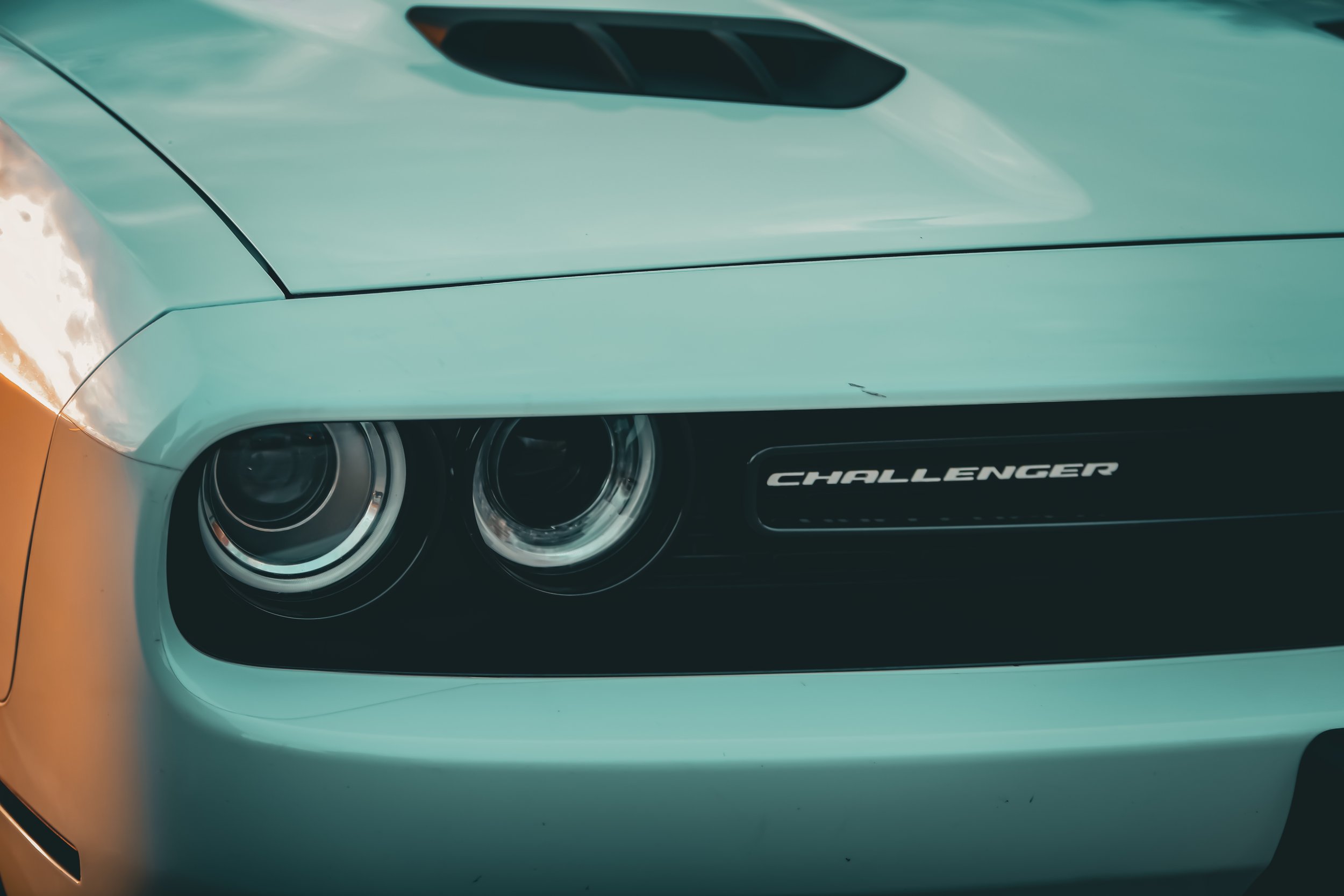 Close-up of the front end of a light green Dodge Challenger, showing the dual headlights and the 'Challenger' badge on the grille.