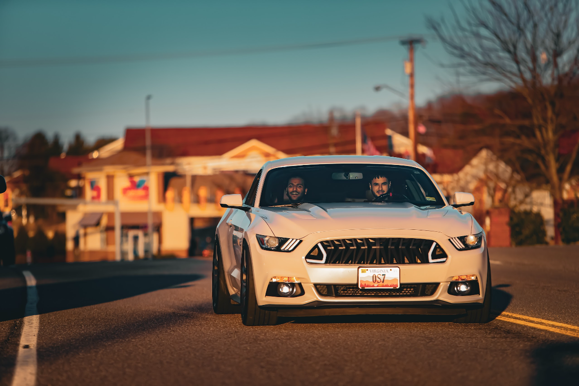 A white sports car driving on a two-lane road with two men inside, one smiling and both looking forward. The background features houses and trees under a sunset sky.