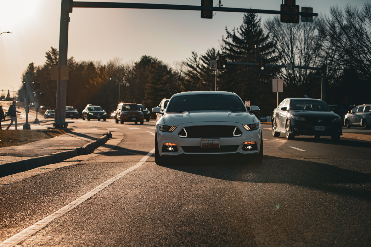 A white sports car driving towards the camera on a city street during sunset, with other vehicles and pedestrians visible in the background.