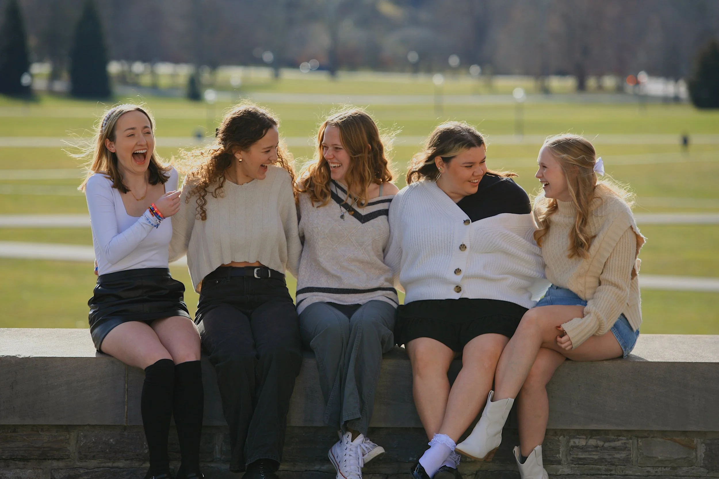 Six young women sitting on a stone wall outdoors, laughing and enjoying each other's company on a sunny day.