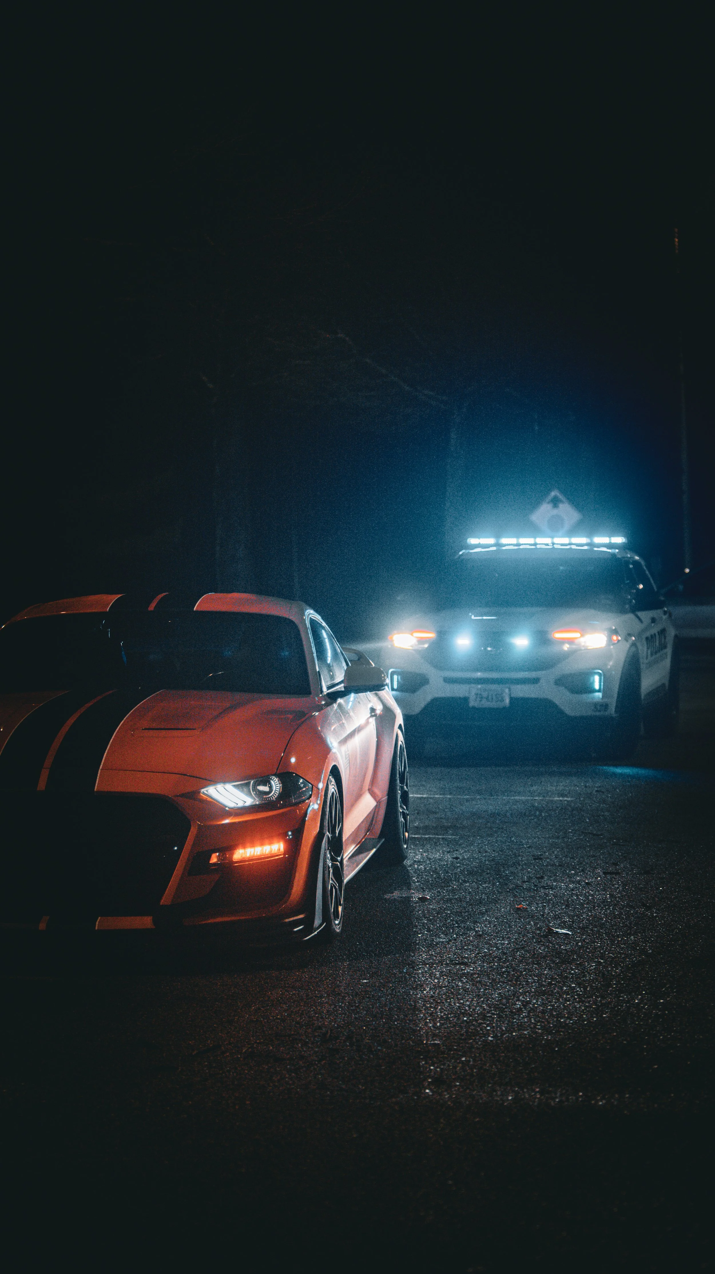 A red sports car with black racing stripes parked on a wet road at night, with a police car with flashing lights behind it.