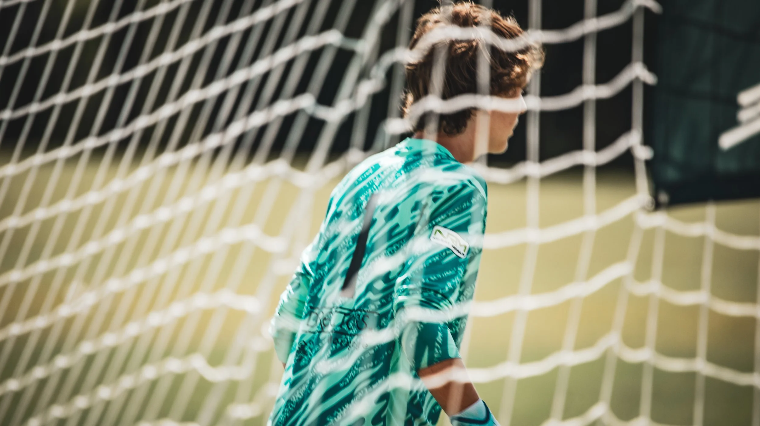 A soccer goalkeeper standing behind the goal net on a sunny field.