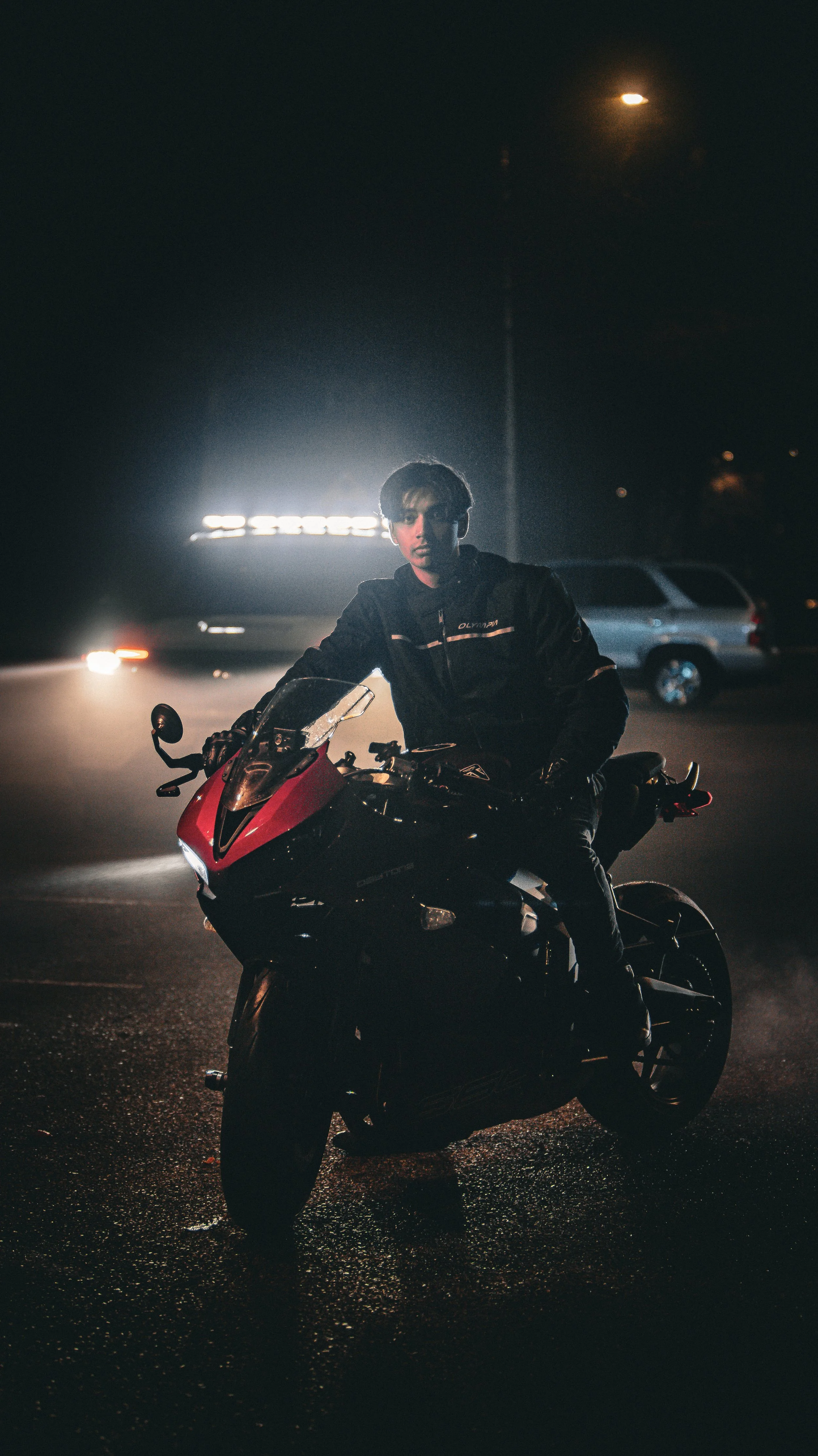 A young man in dark clothing sitting on a red and black motorcycle at night, with street lights and cars visible in the background.