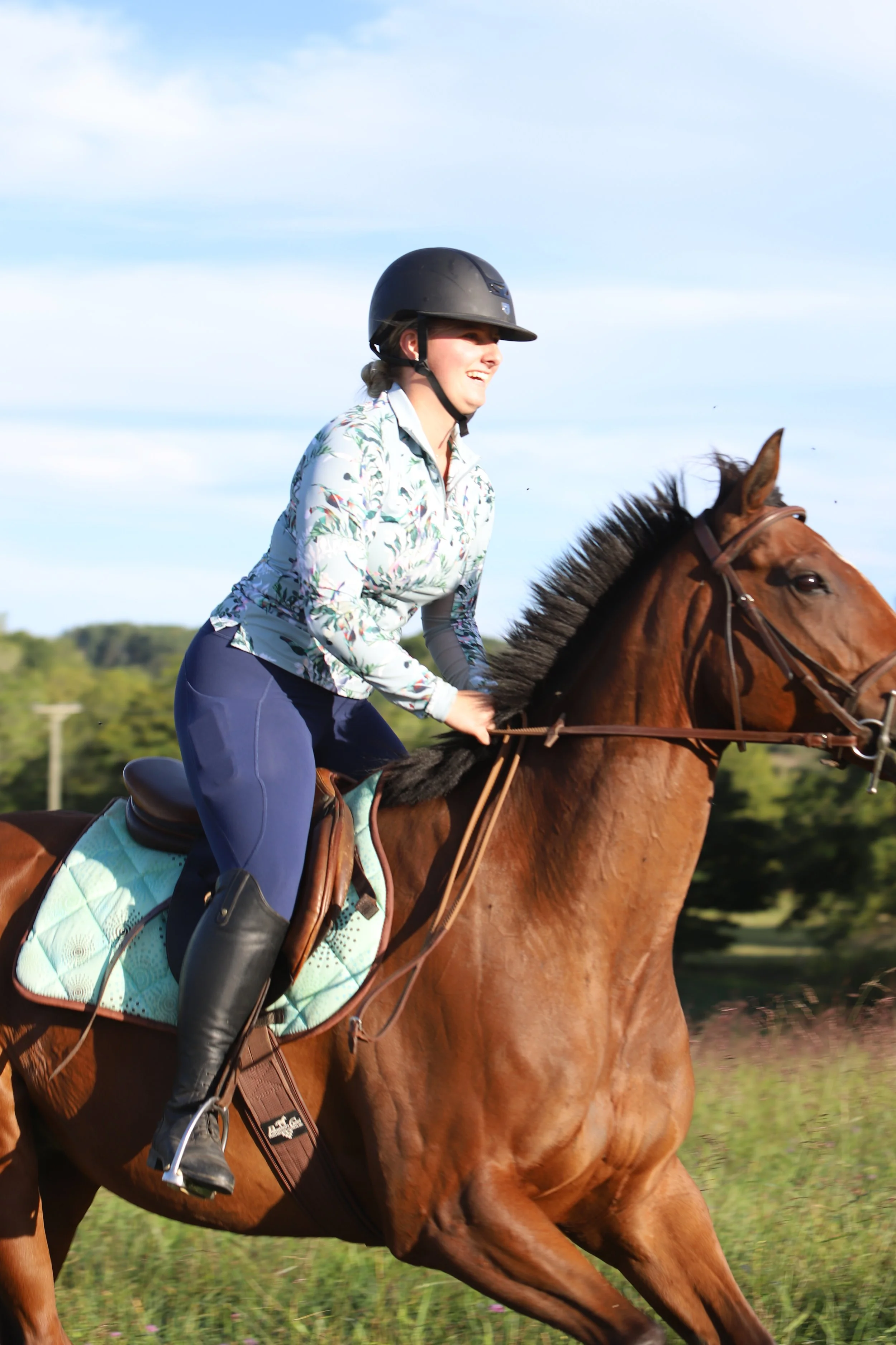 A woman riding a galloping brown horse in a field, wearing a riding helmet, floral jacket, navy riding pants, and tall black riding boots, with a blue sky and green trees in the background.
