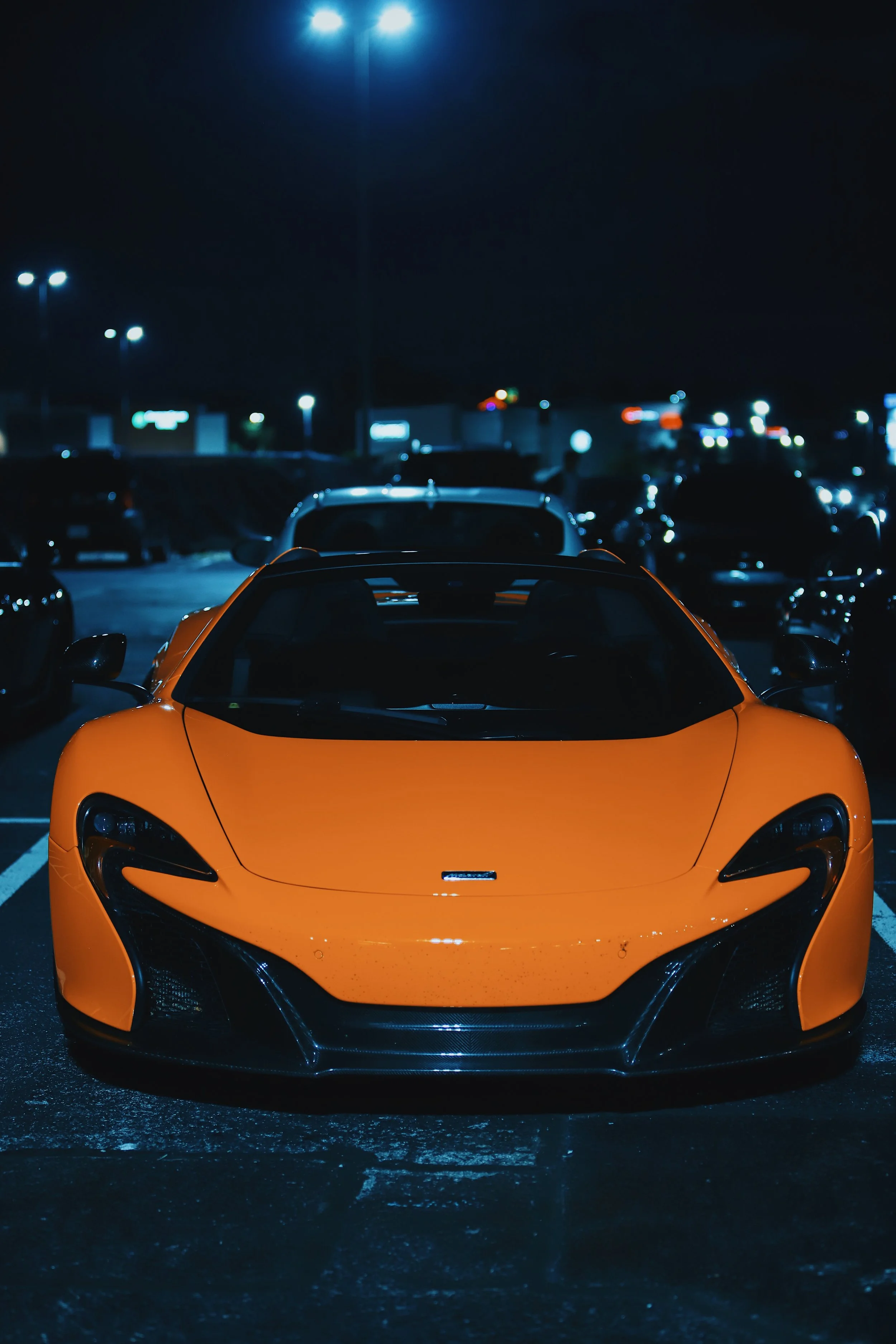 A bright orange sports car parked in a dark parking lot at night, illuminated by streetlights and other cars in the background.
