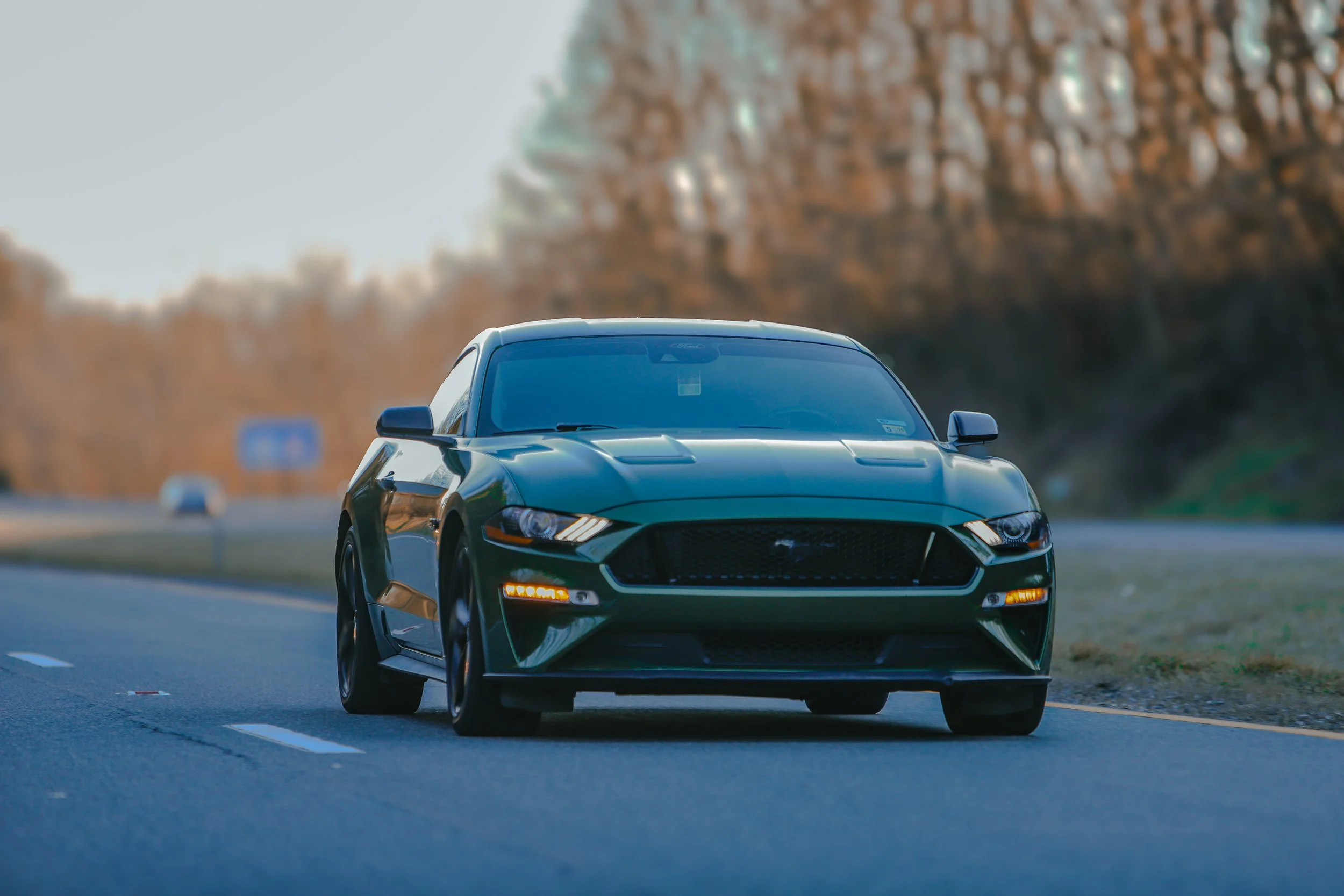 A black Ford Mustang sports car driving on an open road with trees in the background.