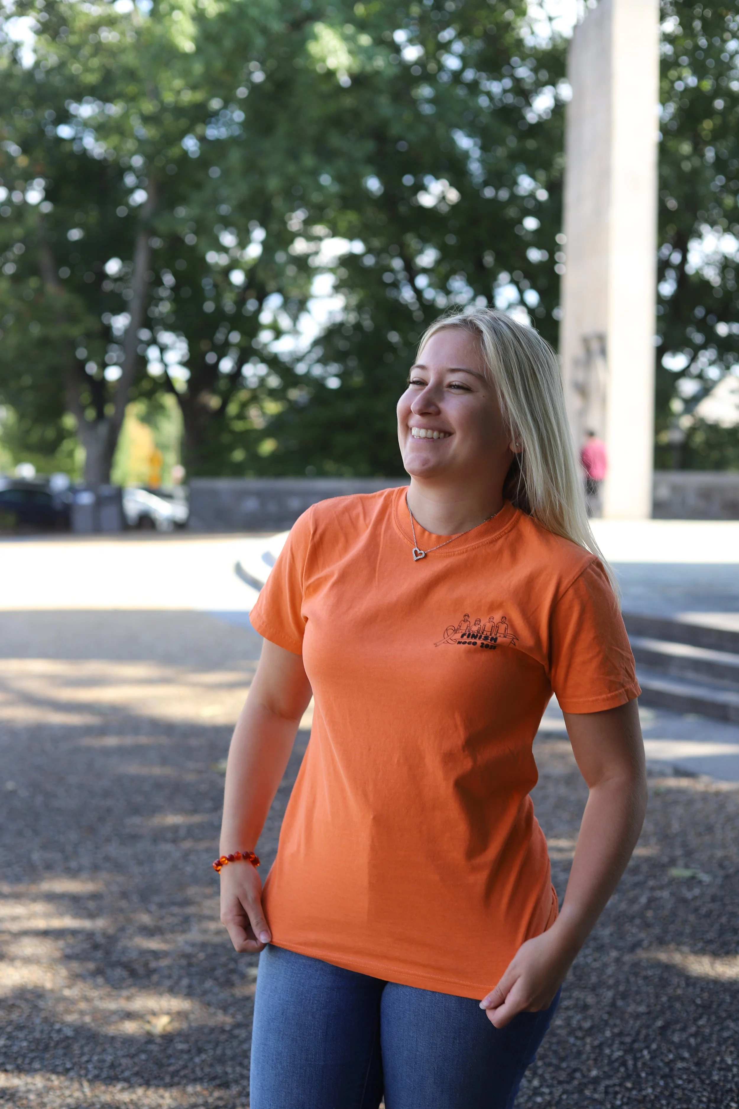 A young woman with blonde hair smiling outdoors on a sunny day, wearing an orange T-shirt and blue jeans, with trees and stairs in the background.