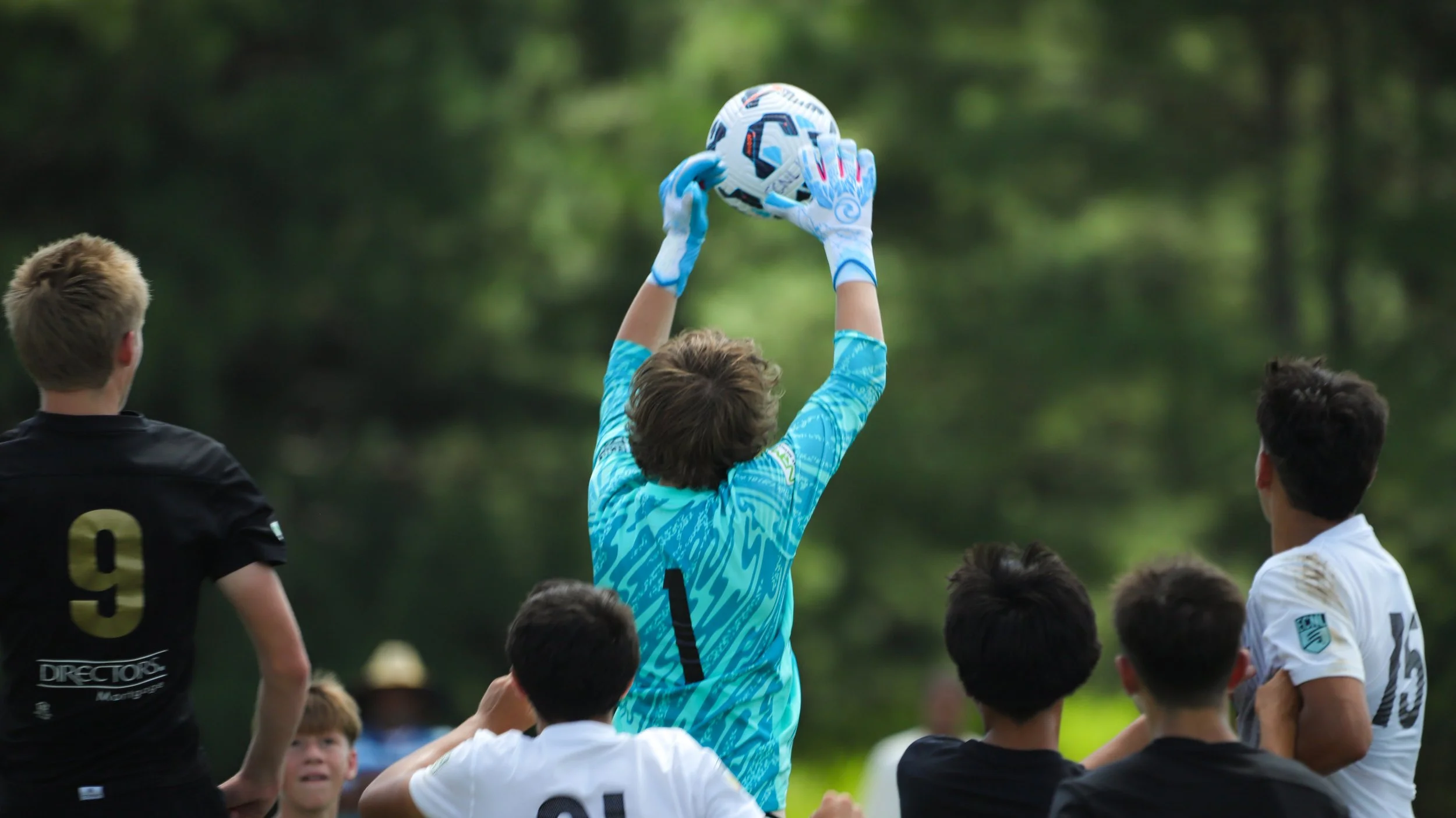 Soccer players on the field during a match, with a goalkeeper in a blue jersey catching the ball. Other players in black and white jerseys are nearby.