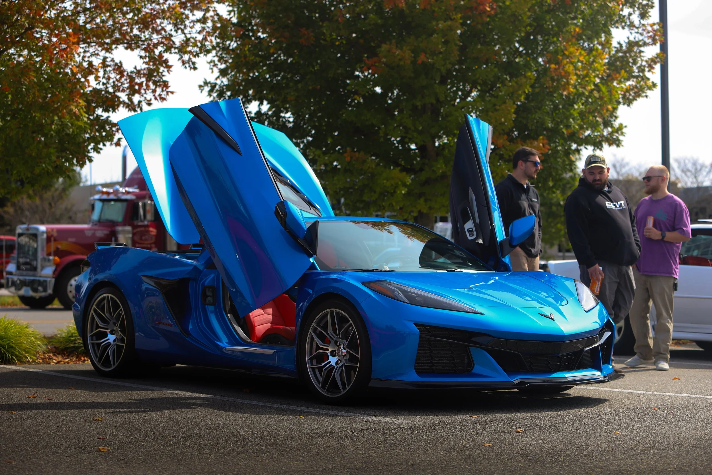 A blue sports car with its doors open, showing red interior, parked outdoors with trees and people nearby.