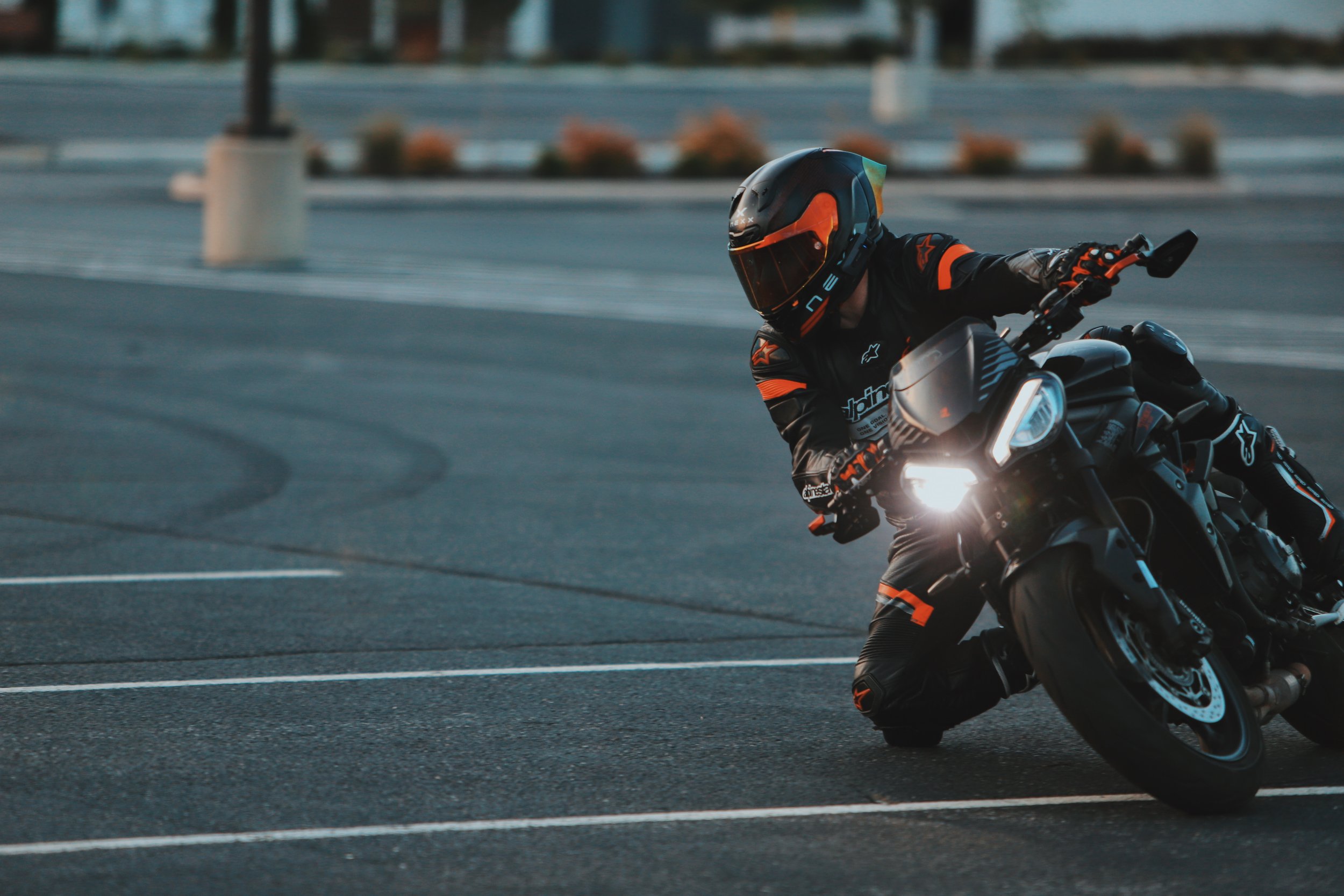 Motorcycle rider in black and orange riding gear on a black motorcycle, leaning into a turn in a parking lot.
