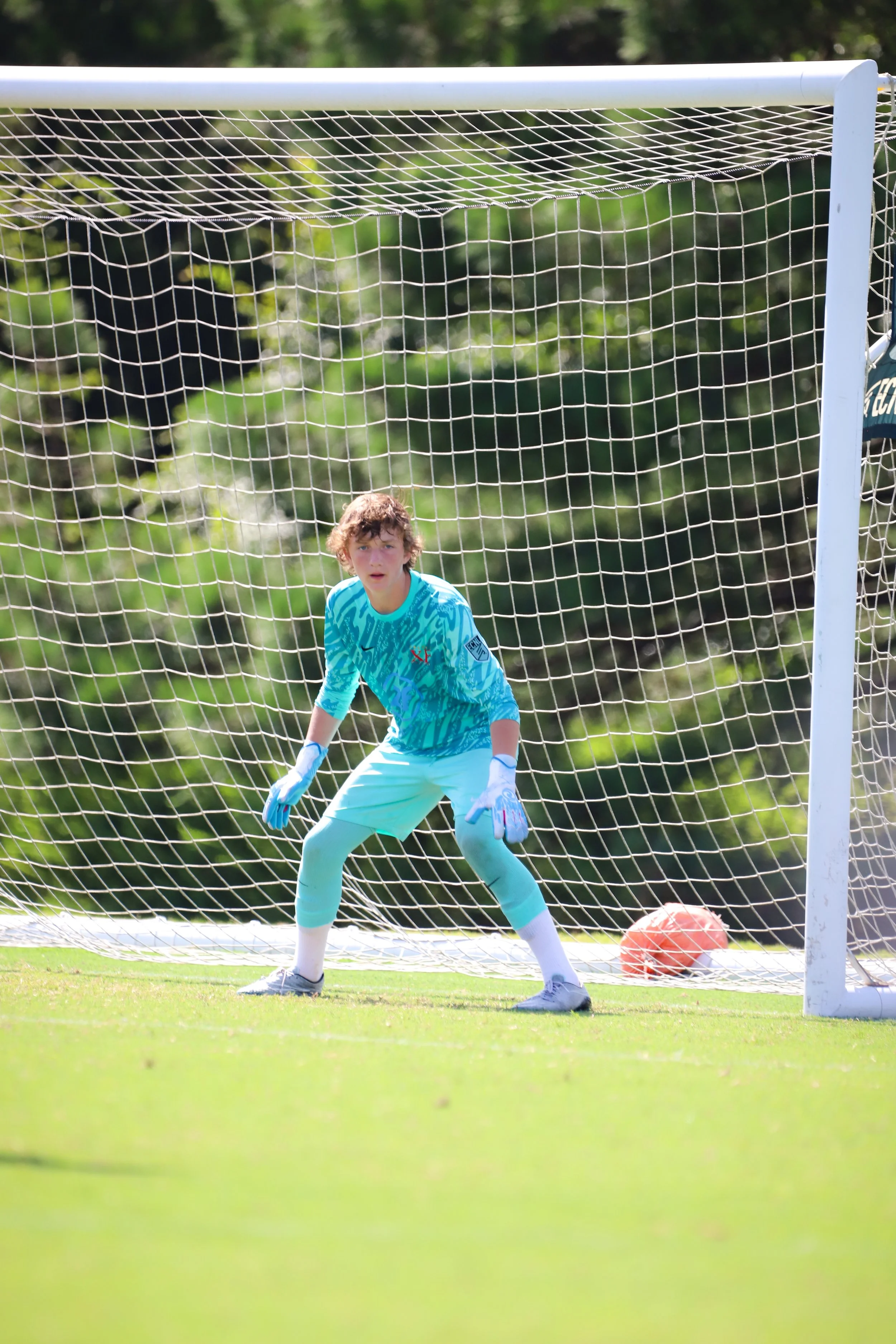 A young soccer goalkeeper in a light blue uniform and gloves stands alert in front of the goal with a soccer ball behind her on the grass field, surrounded by greenery.