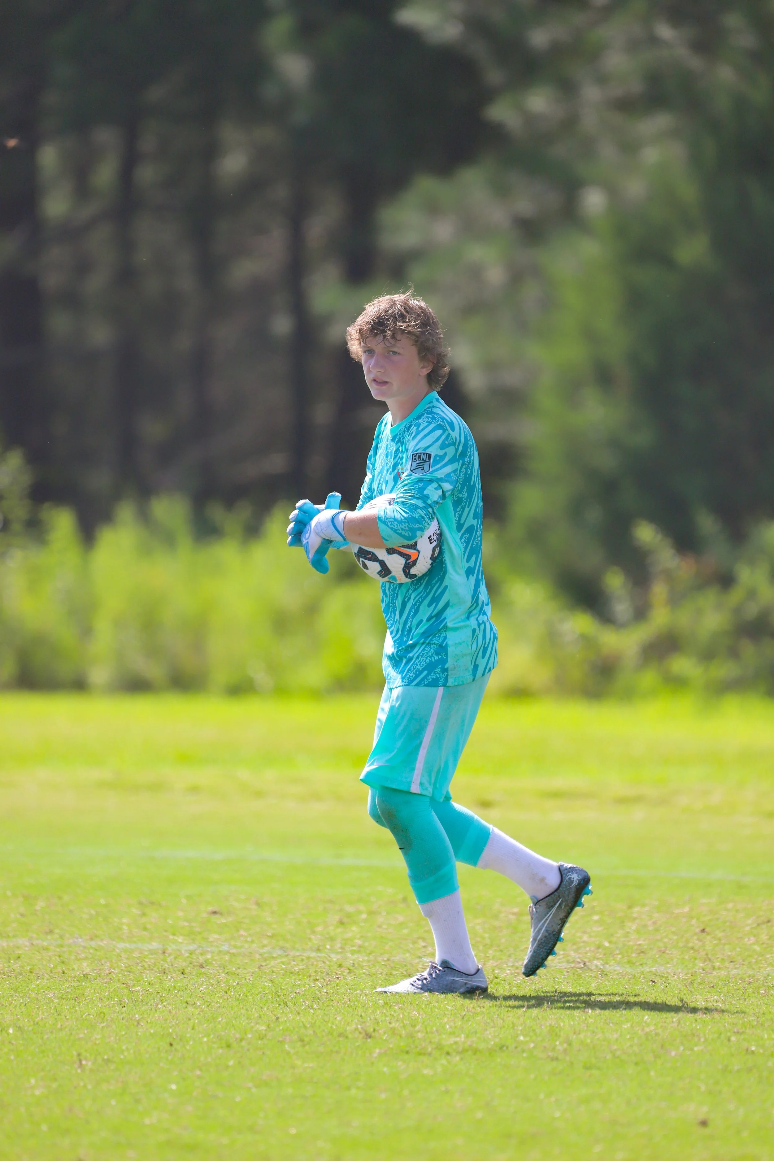 A young soccer goalkeeper in a blue uniform holding a soccer ball on a grassy field.