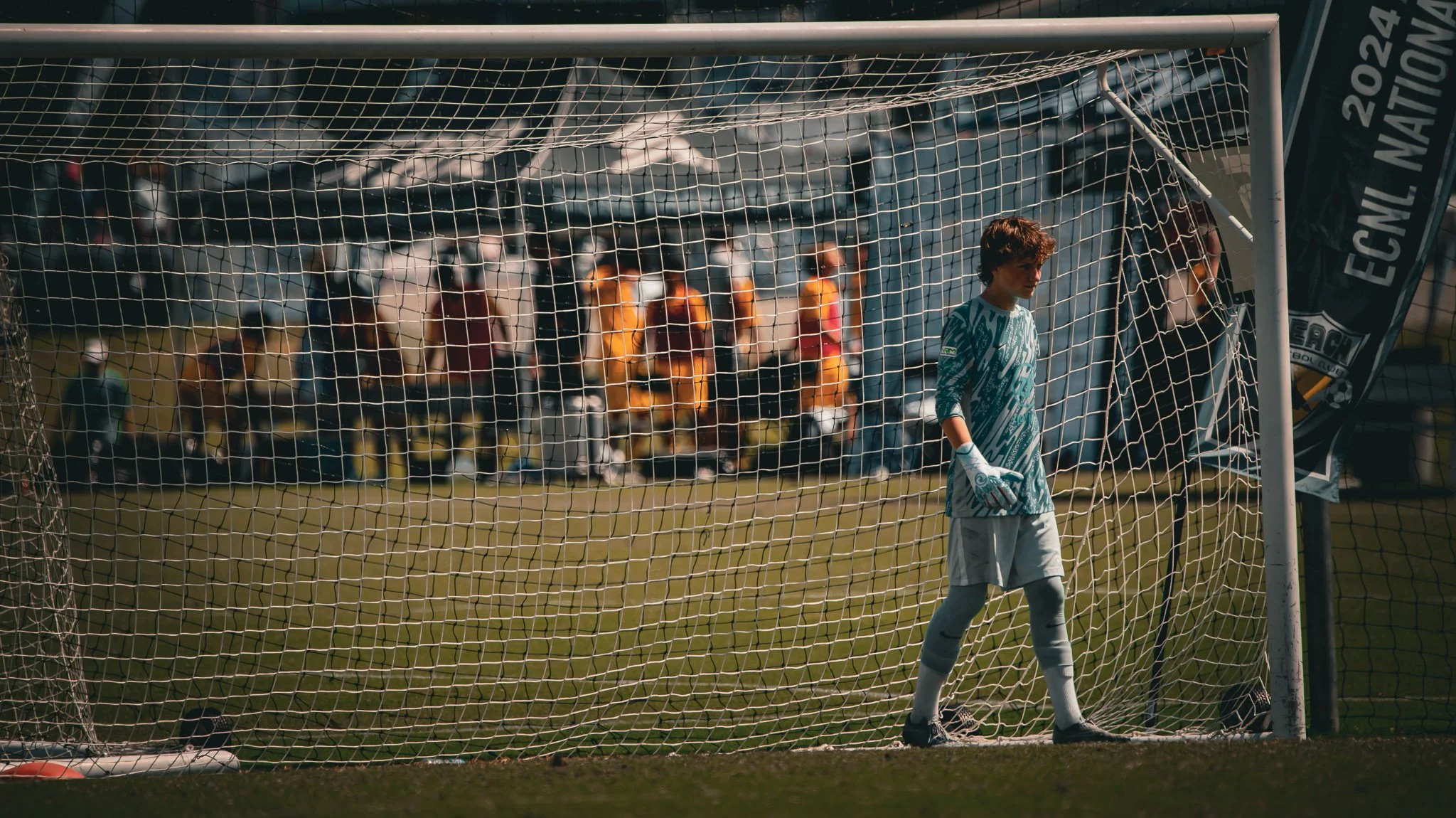 A young male soccer goalkeeper standing inside the goal net during a match, with players and spectators blurred in the background.