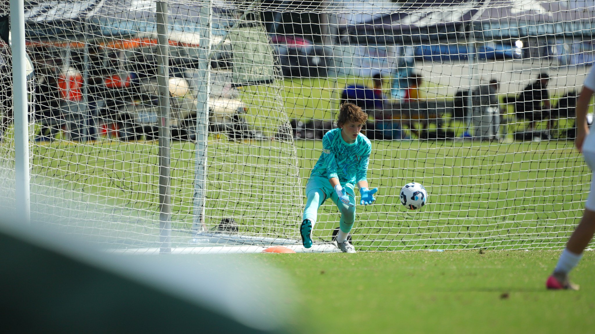 A young soccer goalkeeper in a blue uniform and gloves is diving to save a soccer ball near the goalpost on a grassy field, with spectators and cars in the background.