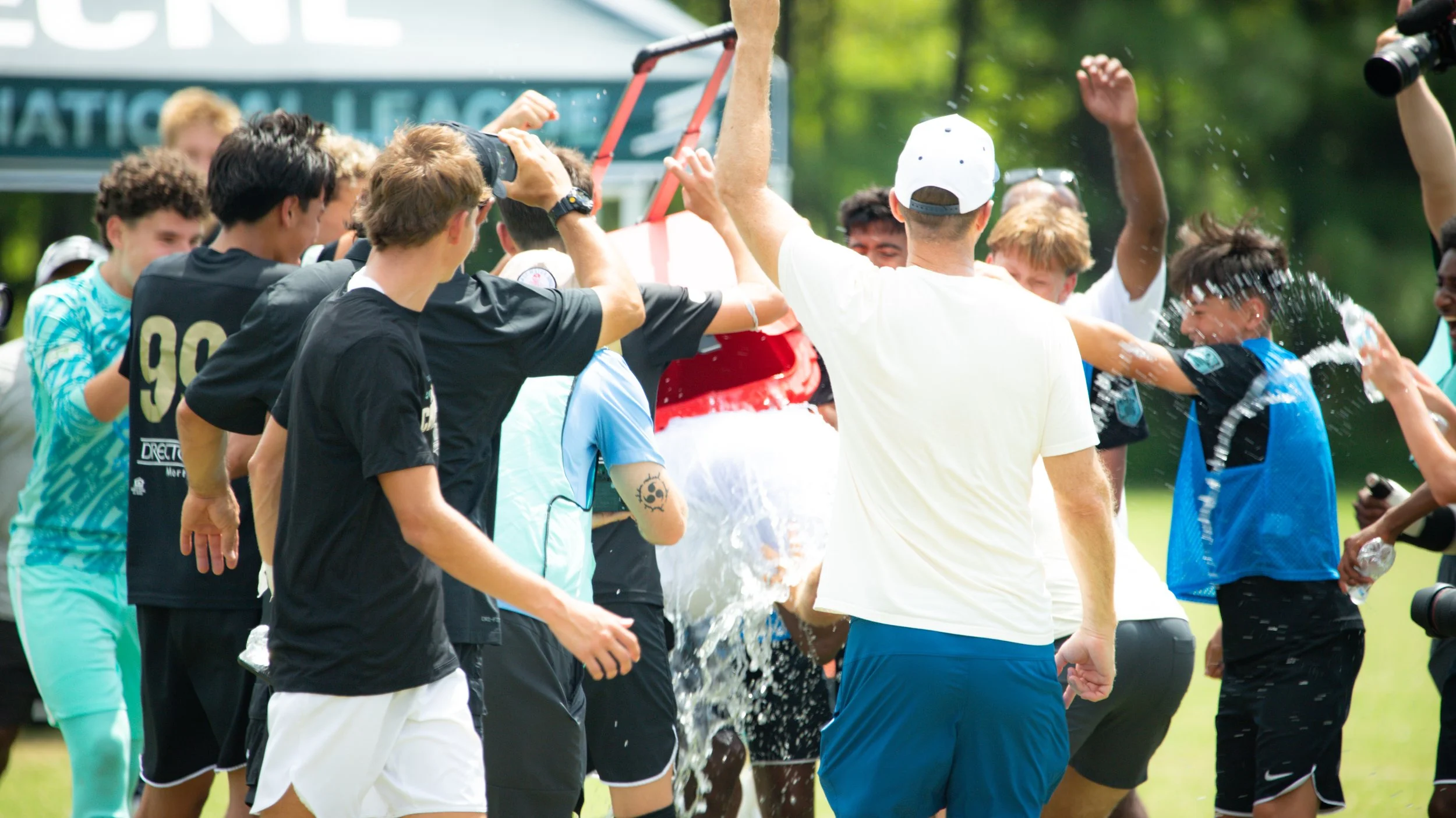 Group of young people celebrating outdoors, with some splashing and pouring water on each other, in a lively scene at what appears to be a sports event or gathering.