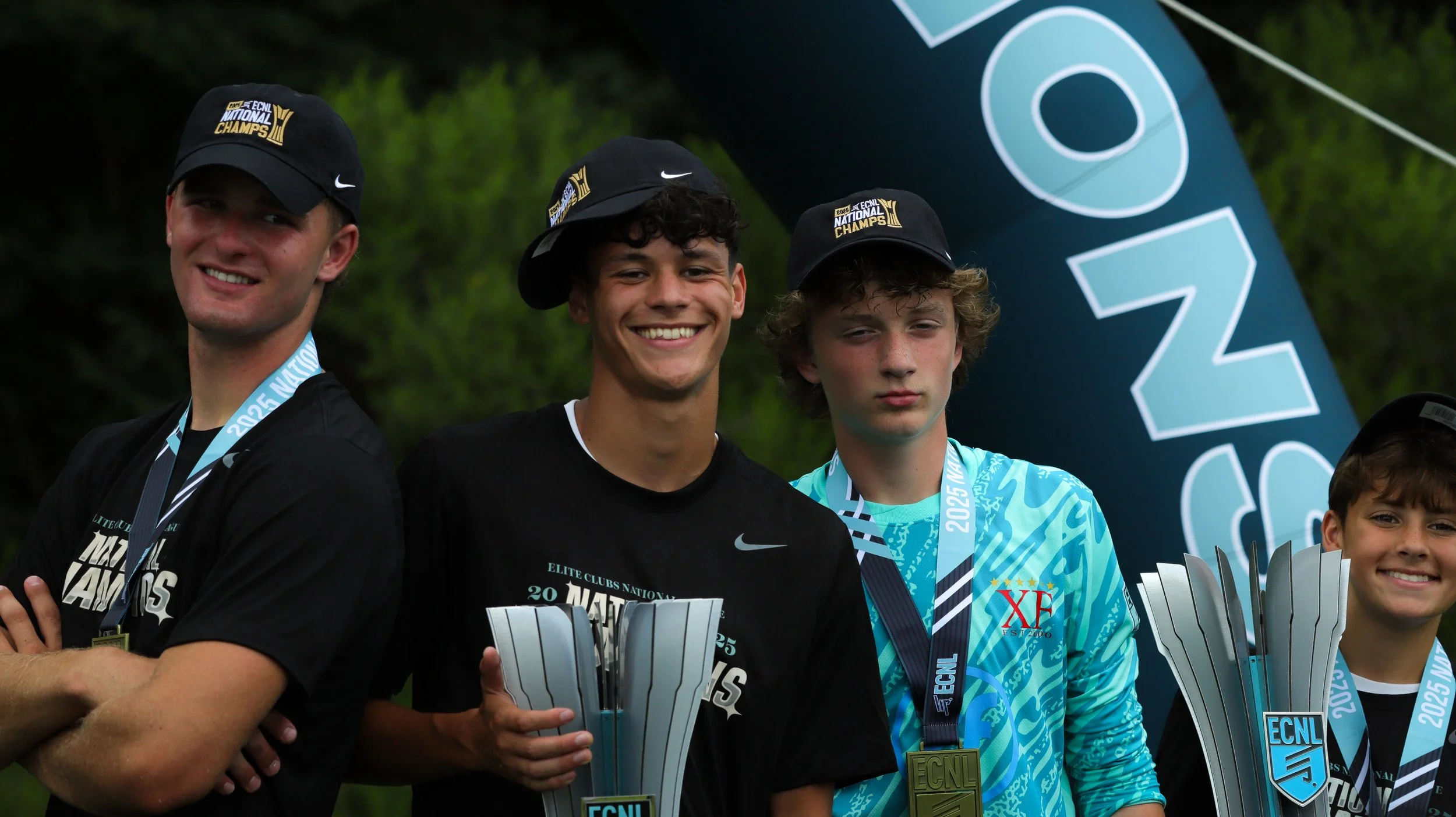 Young male athletes celebrating with trophies and medals at an award ceremony outdoors, wearing black caps and sports shirts, with a large ECNL banner in the background.