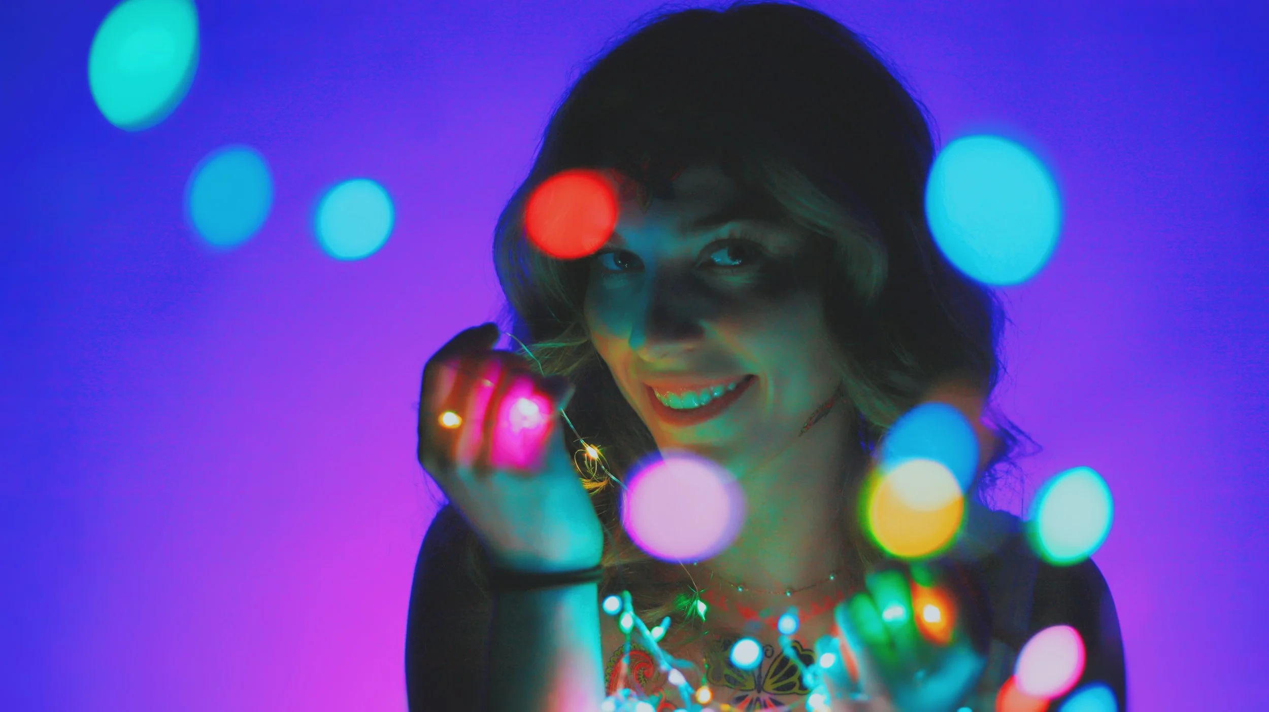 A woman smiling while holding colorful string lights in front of a purple background, with a bokeh effect of multicolored lights surrounding her.
