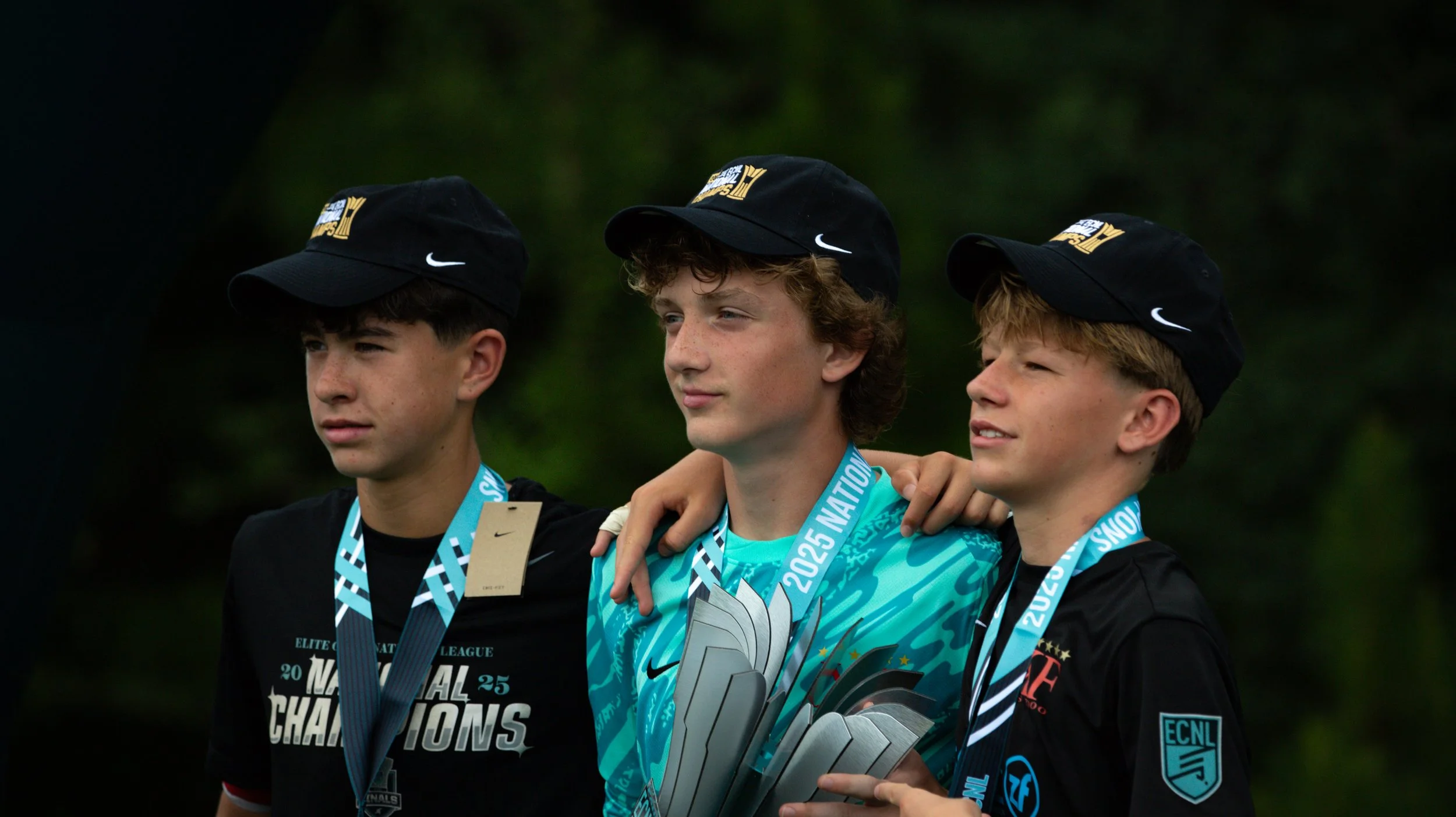 Three young boys wearing black caps and black shirts with medals around their necks, standing on a podium with a dark, blurry outdoor background, celebrating a sports victory.
