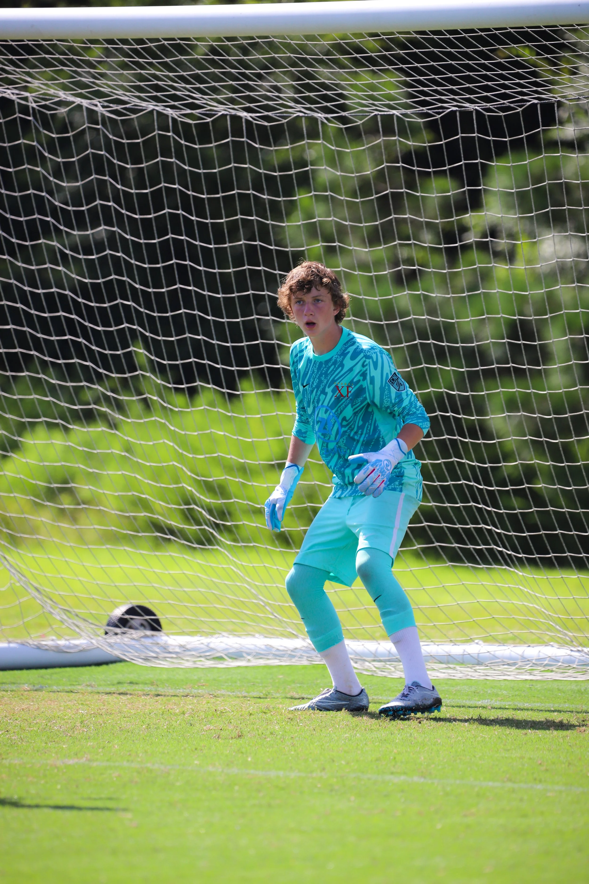 A young male soccer goalkeeper standing in front of a goal net during a match, wearing a turquoise goalkeeper uniform and gloves on a grassy field.