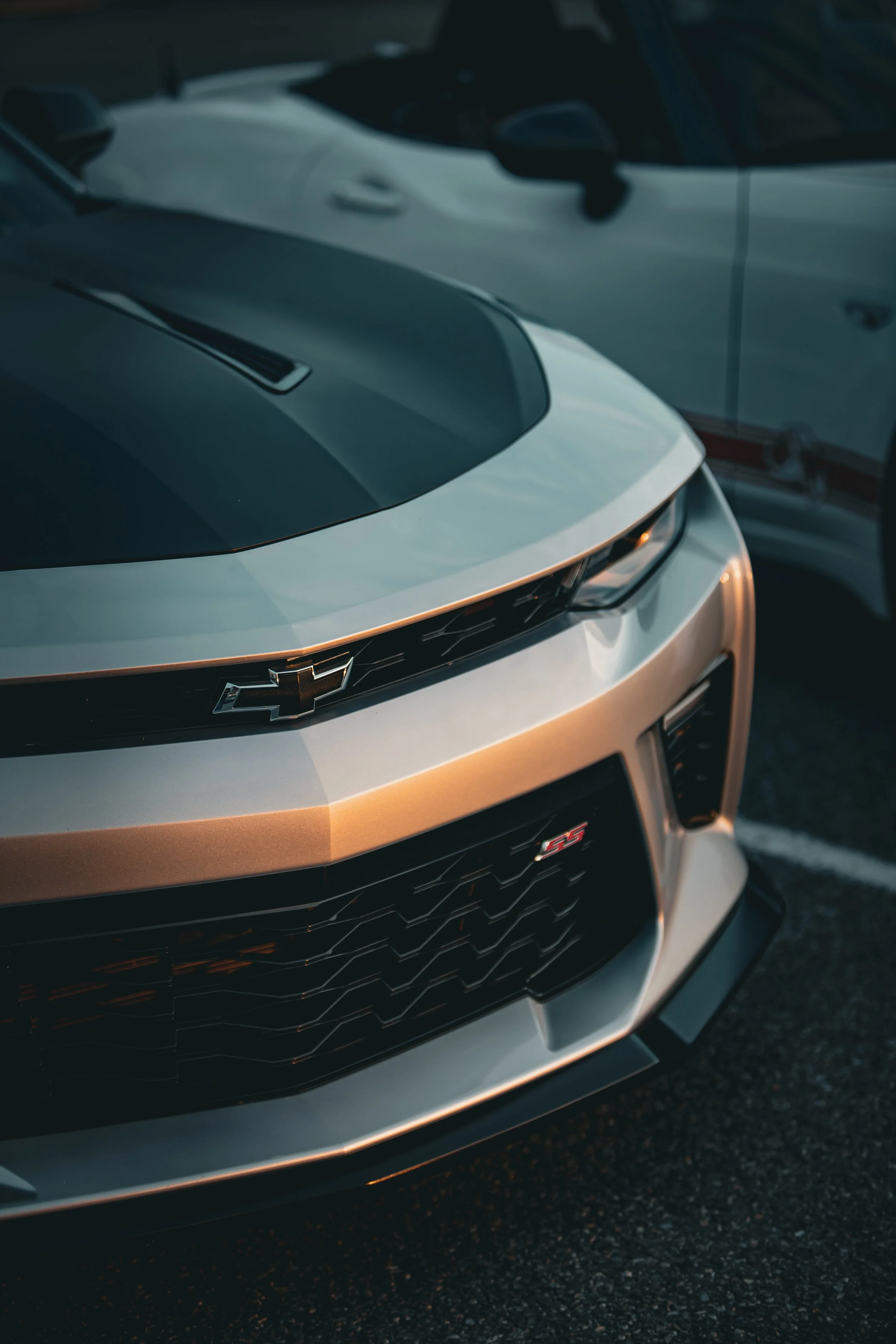 Close-up of the front of a silver Chevrolet Camaro SS car parked on a street at dusk, with a white vehicle in the background.