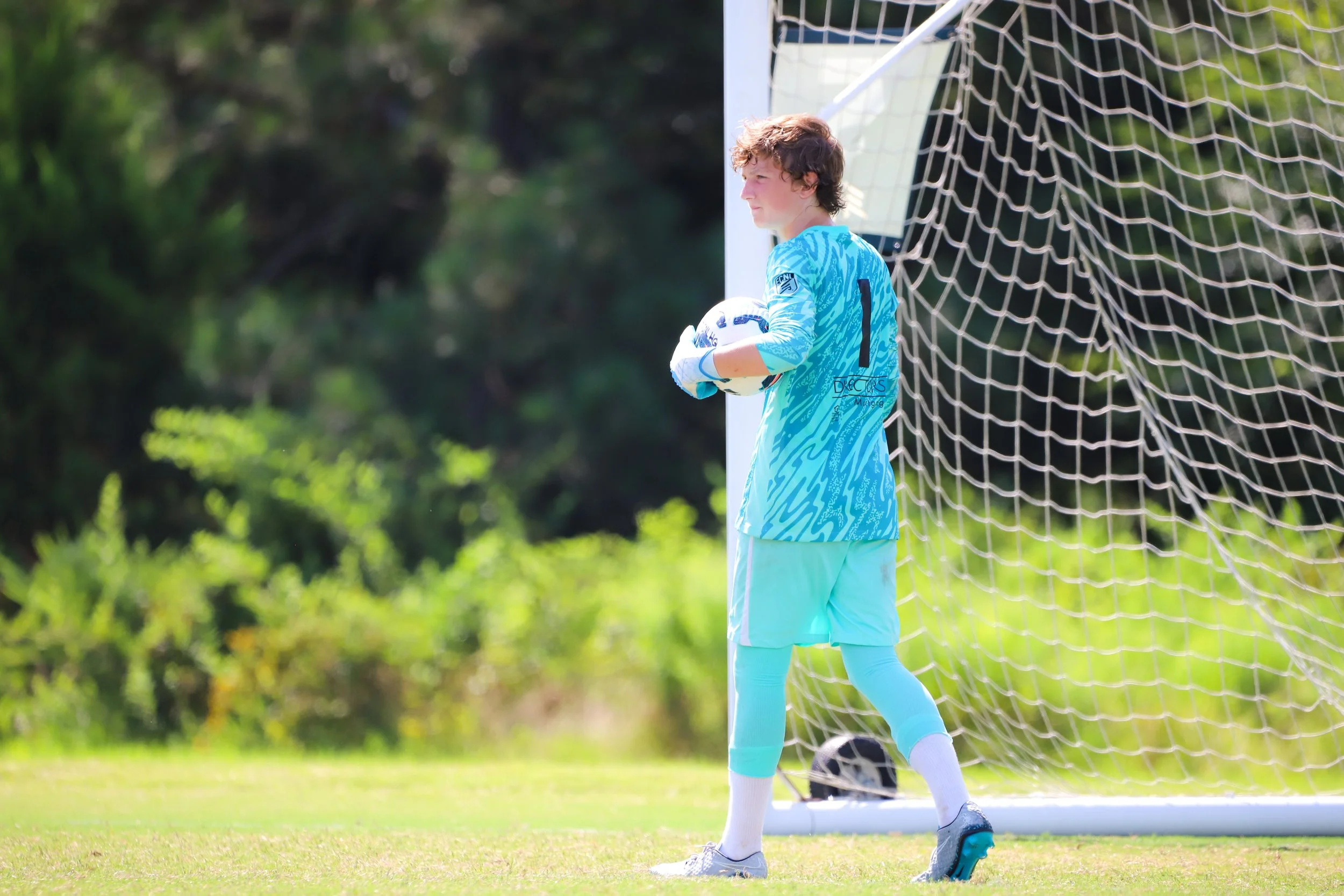 Young male soccer goalkeeper standing on field holding soccer ball, wearing blue uniform and gloves, near goalpost with net, lush greenery in background.