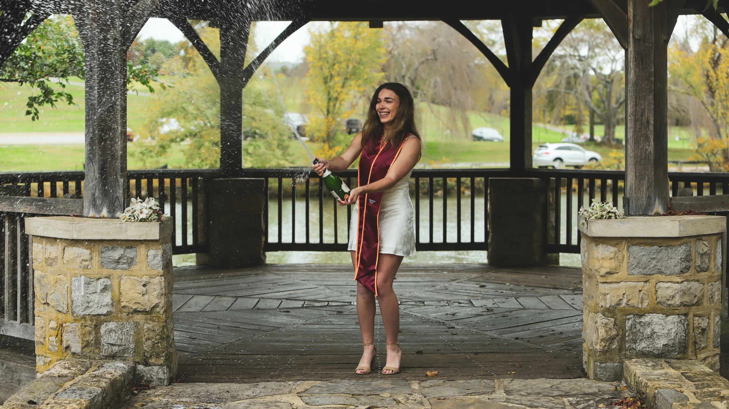 A young woman in a white dress and heels smiling and celebrating as she opens a bottle of champagne, causing it to spray. She is standing on a wooden gazebo with a lake, trees, and parked cars in the background.