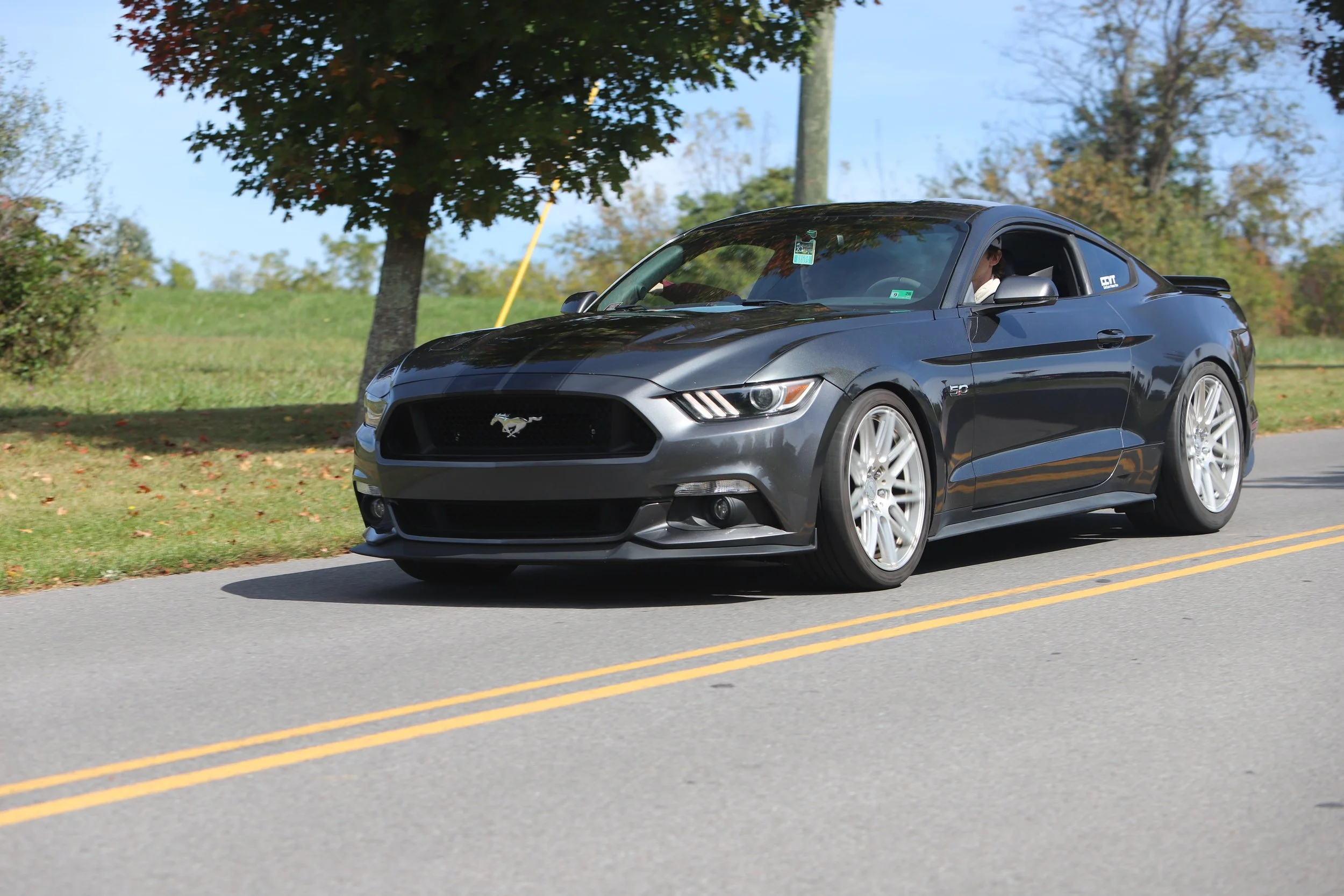 A black Ford Mustang driving on a two-lane road with greenery and trees in the background.