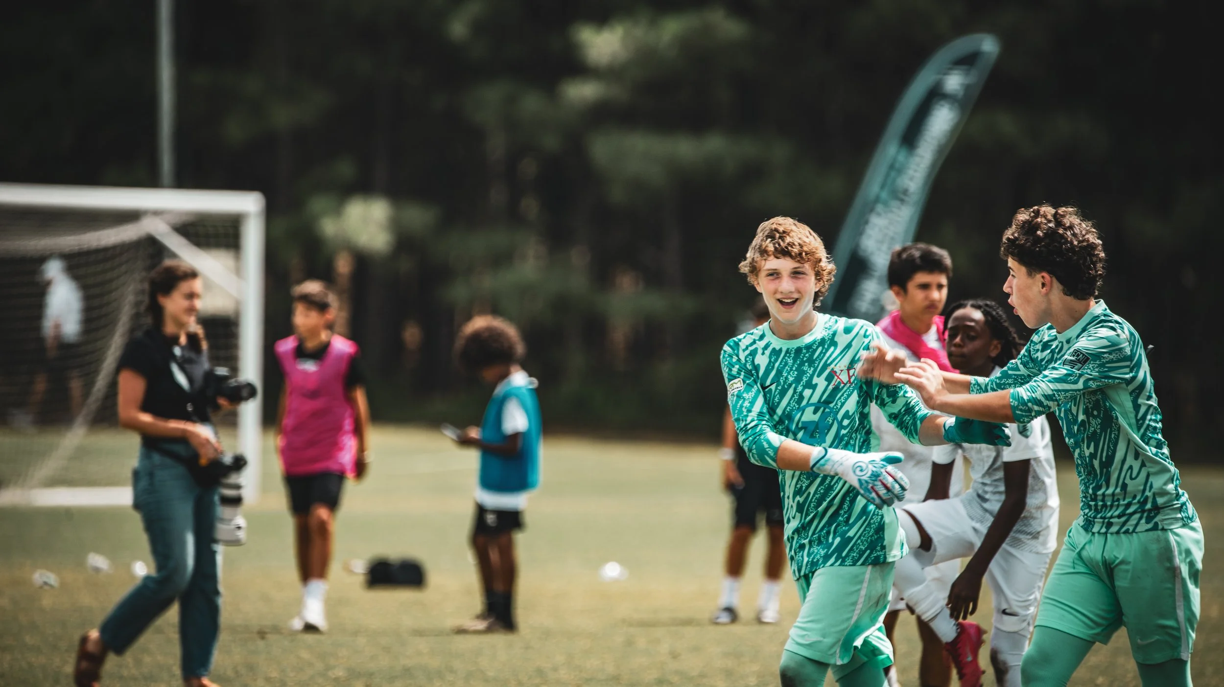 A group of children playing soccer on a field, with a female photographer holding a camera nearby, and a goalpost in the background.