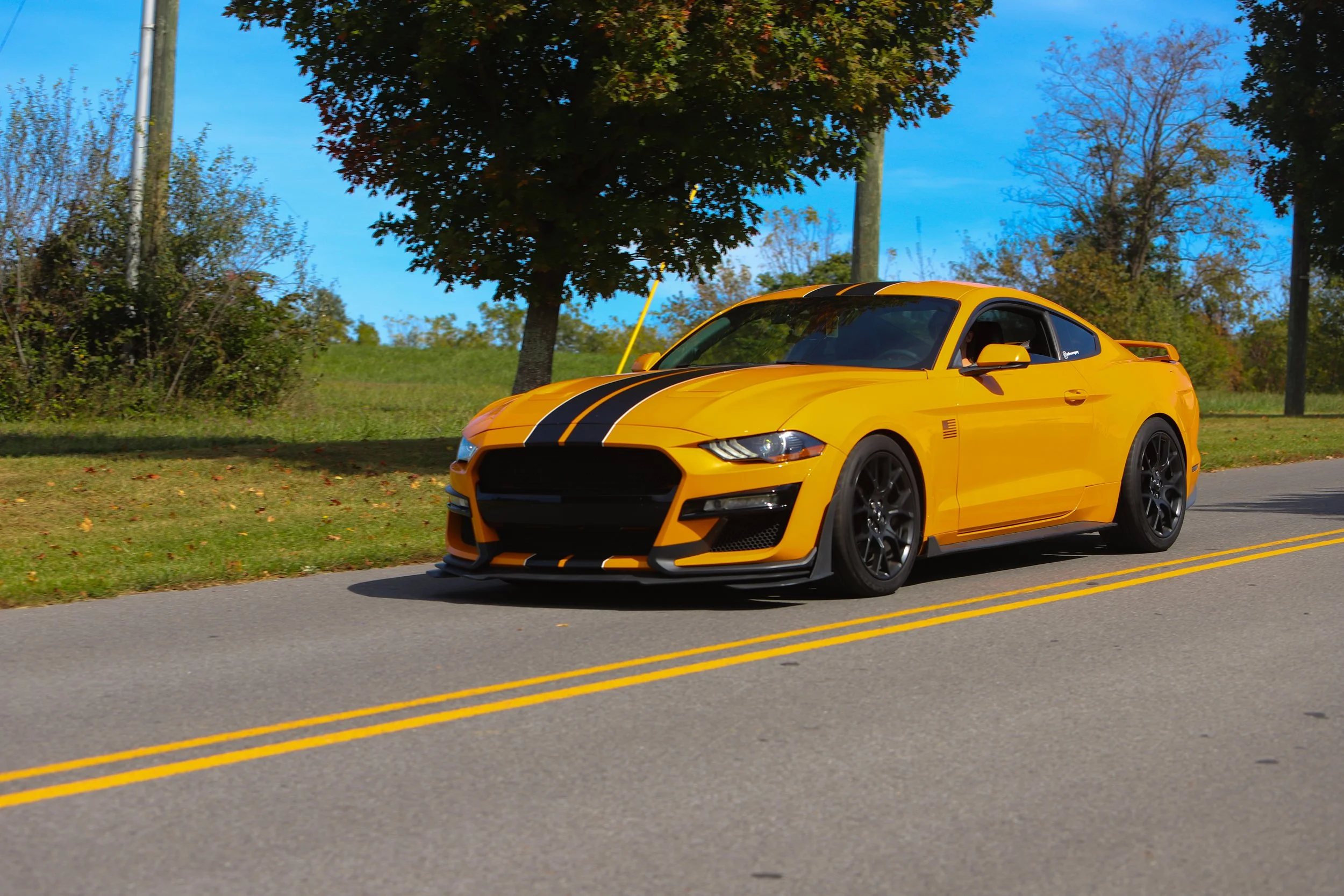 Yellow sports car with black racing stripes driving on a two-lane road with trees and grass on the side, under a partly cloudy sky.
