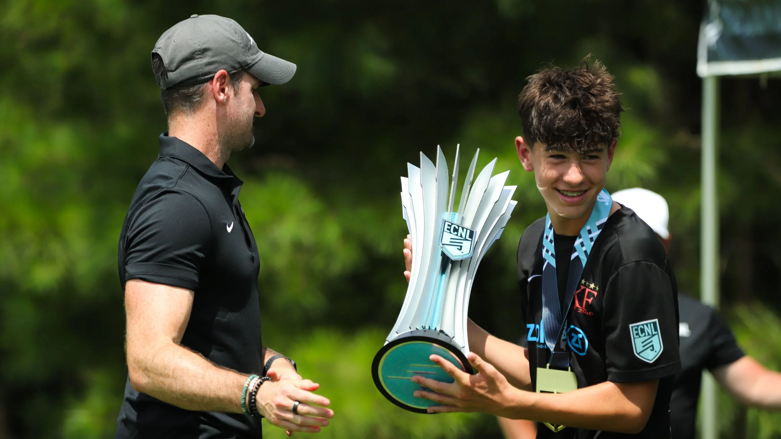 A young boy receiving a trophy from an adult man during an outdoor event, with trees in the background.