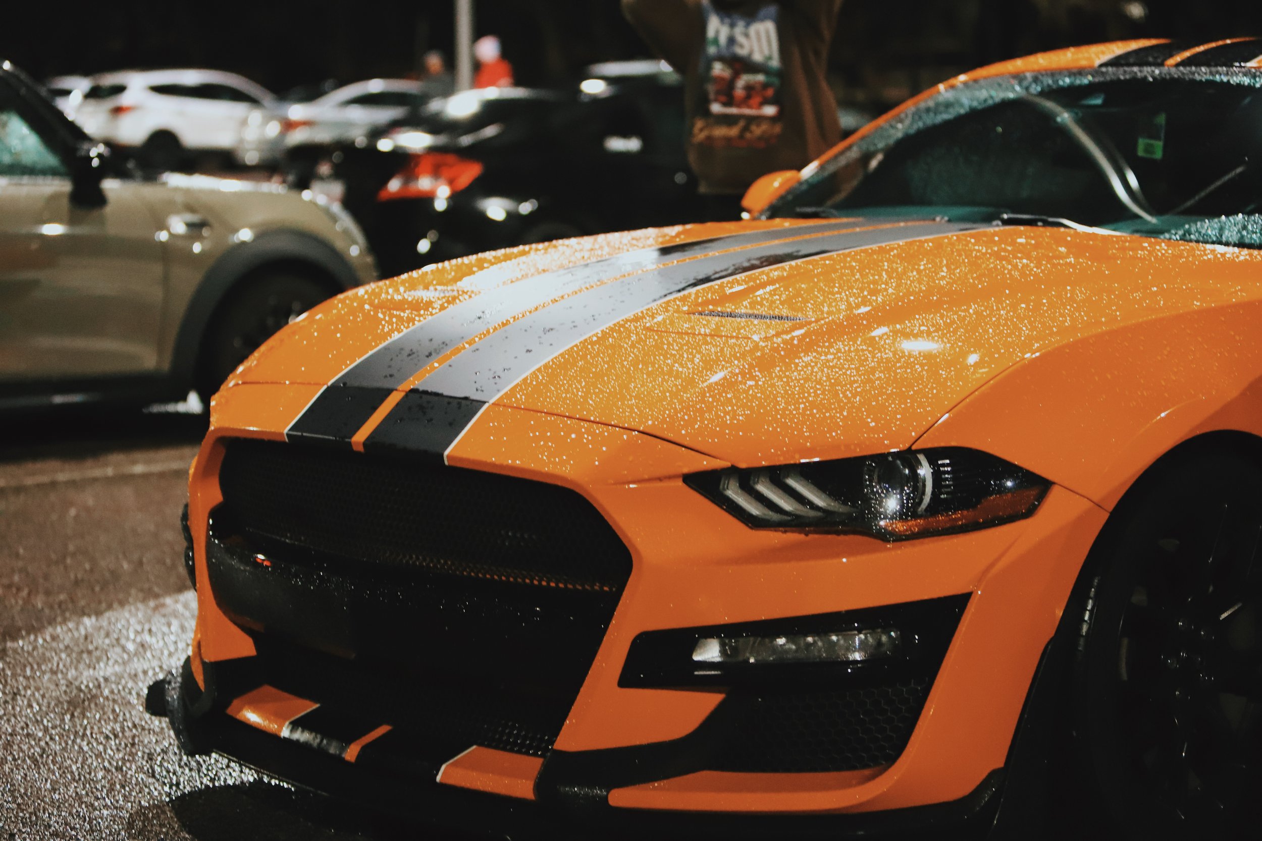 An orange sports car with black racing stripes parked at night, with raindrops on its surface and other cars in the background.