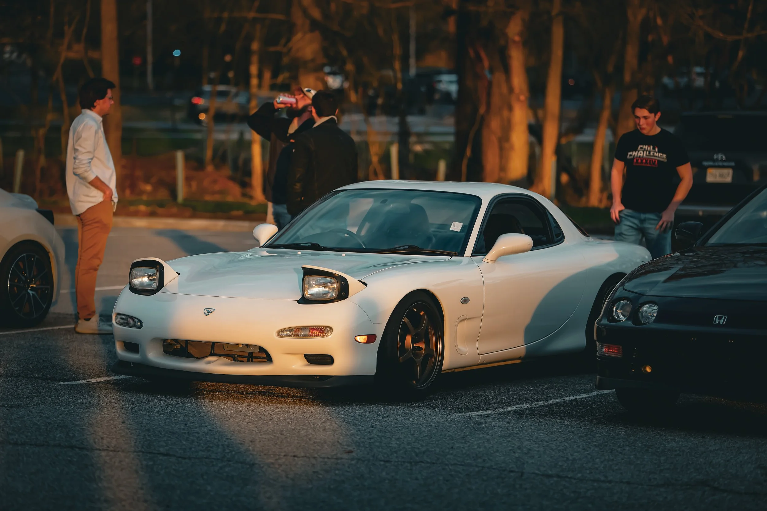A white Mazda RX-7 sports car parked in a lot at sunset, with five people standing around it, some talking and one drinking from a can.