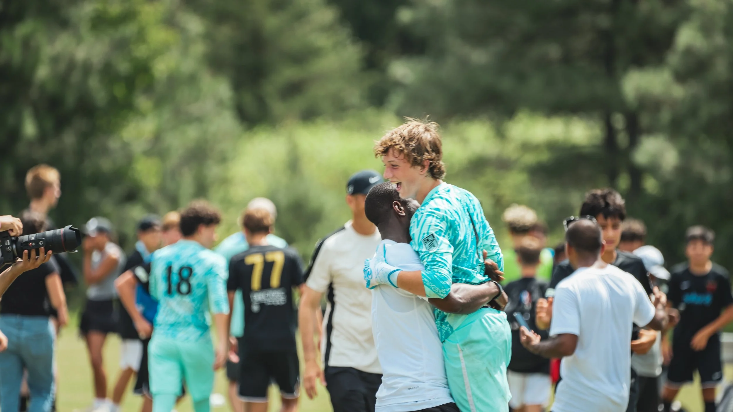 Two soccer players, one in a white jersey and the other in a blue goalkeeper uniform, hugging and celebrating on a soccer field with other players and spectators in the background.