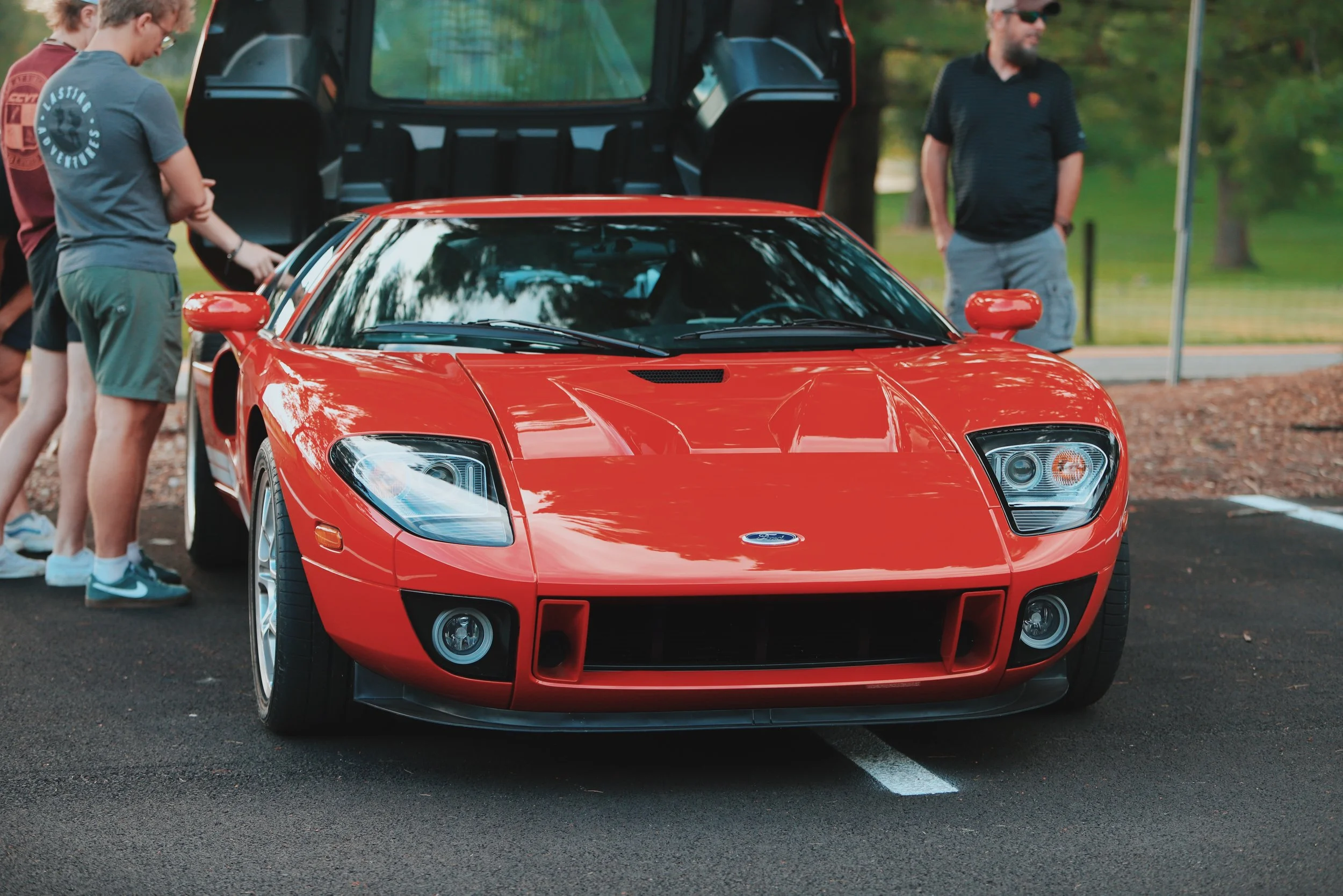 Red Ford sports car parked outdoors with a group of people gathered around it, including a woman in a gray shirt and green shorts, and a man in a black shirt with sunglasses.