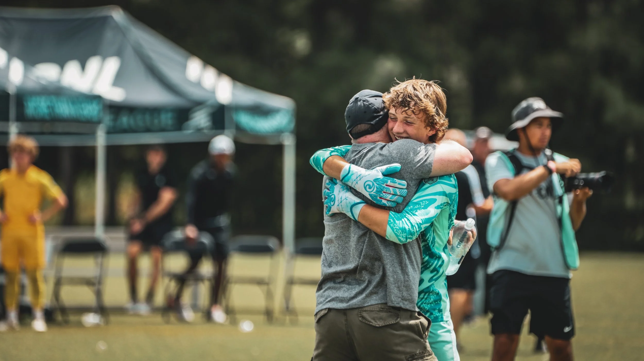 Two people hugging at an outdoor event with a tent and a photographer in the background.