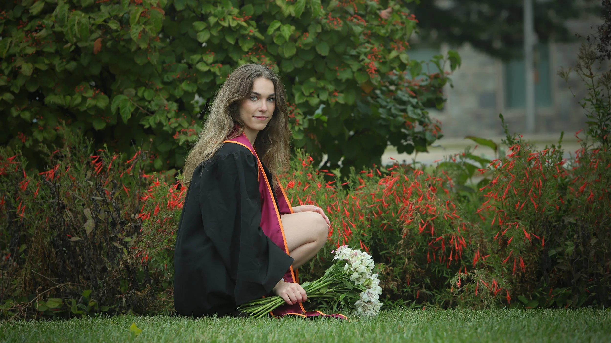 A young woman in a graduation cap and gown kneeling on a grass lawn, holding a bouquet of white flowers, surrounded by lush green and red flowering bushes.
