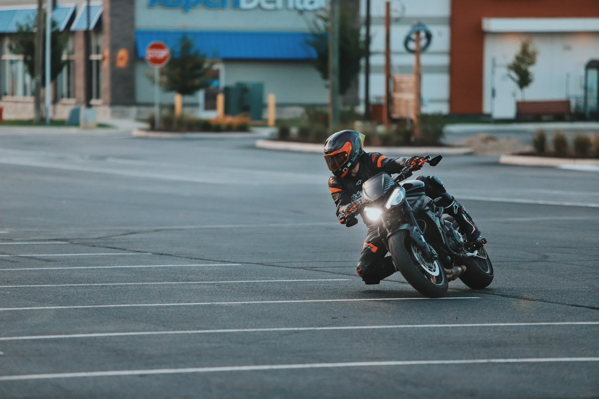 Motorcycle rider wearing a helmet and racing gear leaning into a turn on an empty parking lot outside.