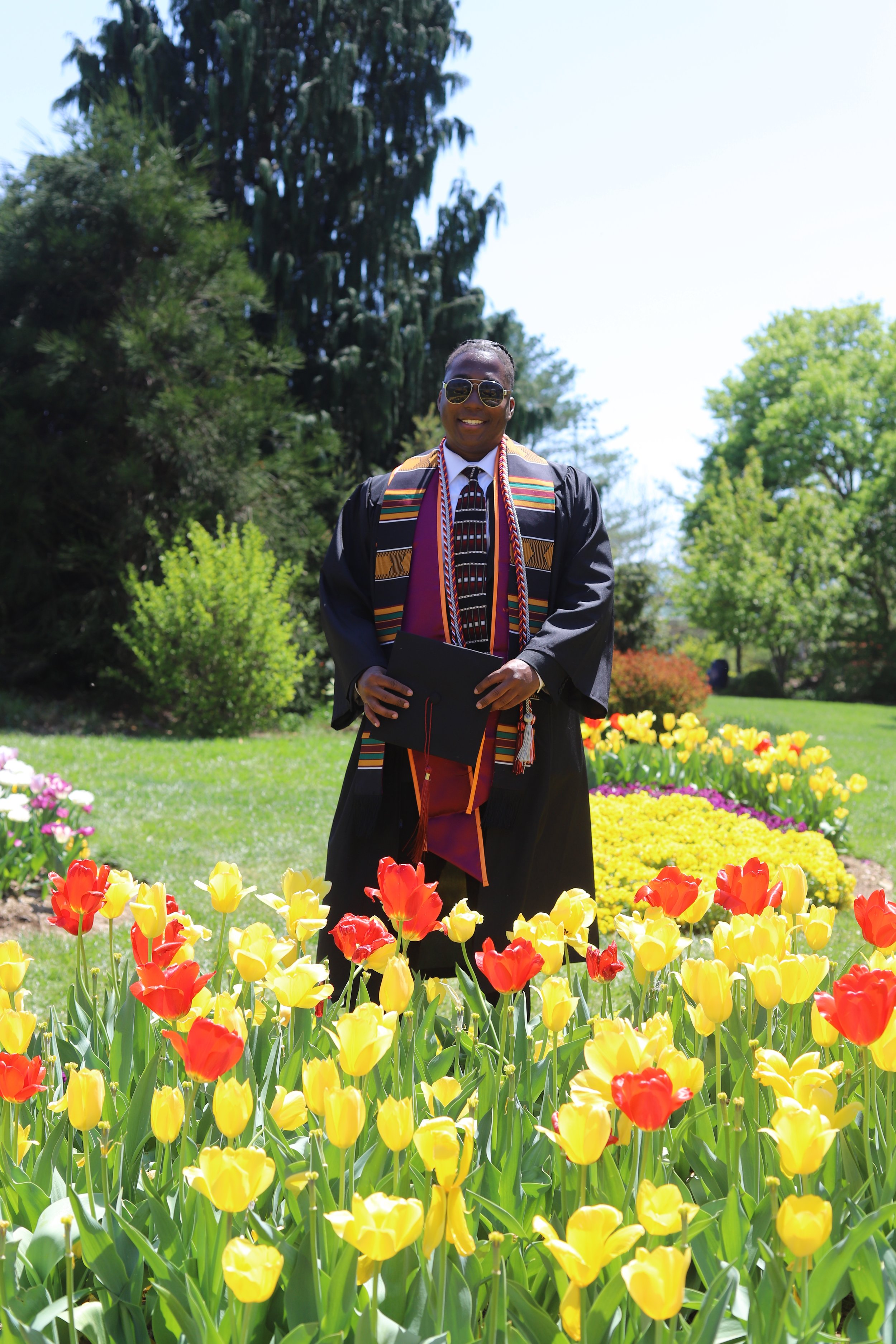 Graduate in cap and gown holding diploma, standing among colorful tulip flowers in a garden with trees.