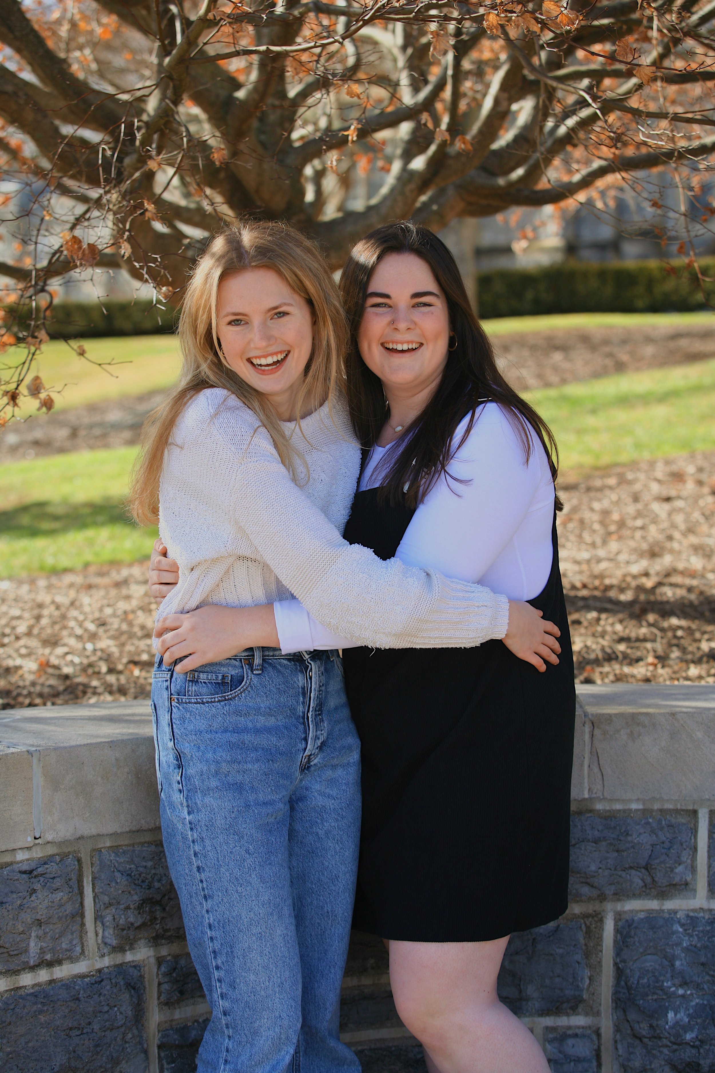 Two smiling young women hugging each other outdoors in front of a tree with autumn leaves.