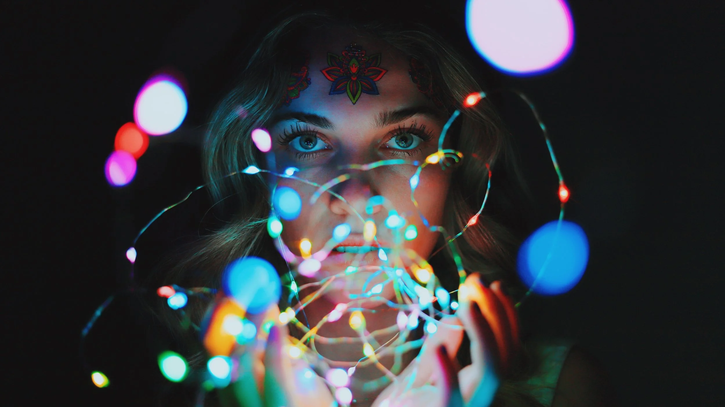 A woman with blue eyes and face tattoos holding colorful string lights, with the lights creating a vibrant glow on her face in a dark background.