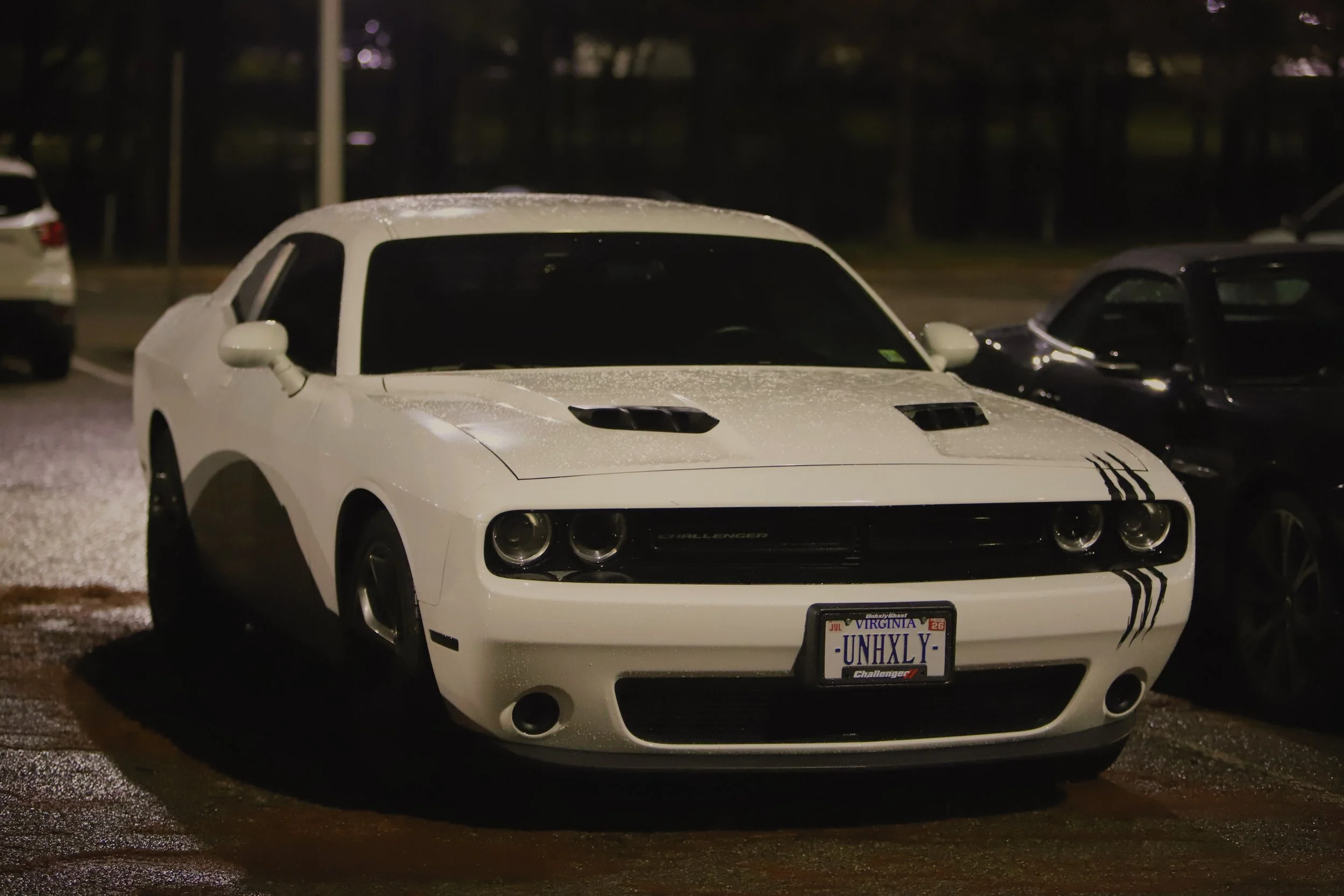White Dodge Challenger sports car parked in a dimly lit parking lot at night, with raindrops on its surface and black stripes on the front fender.