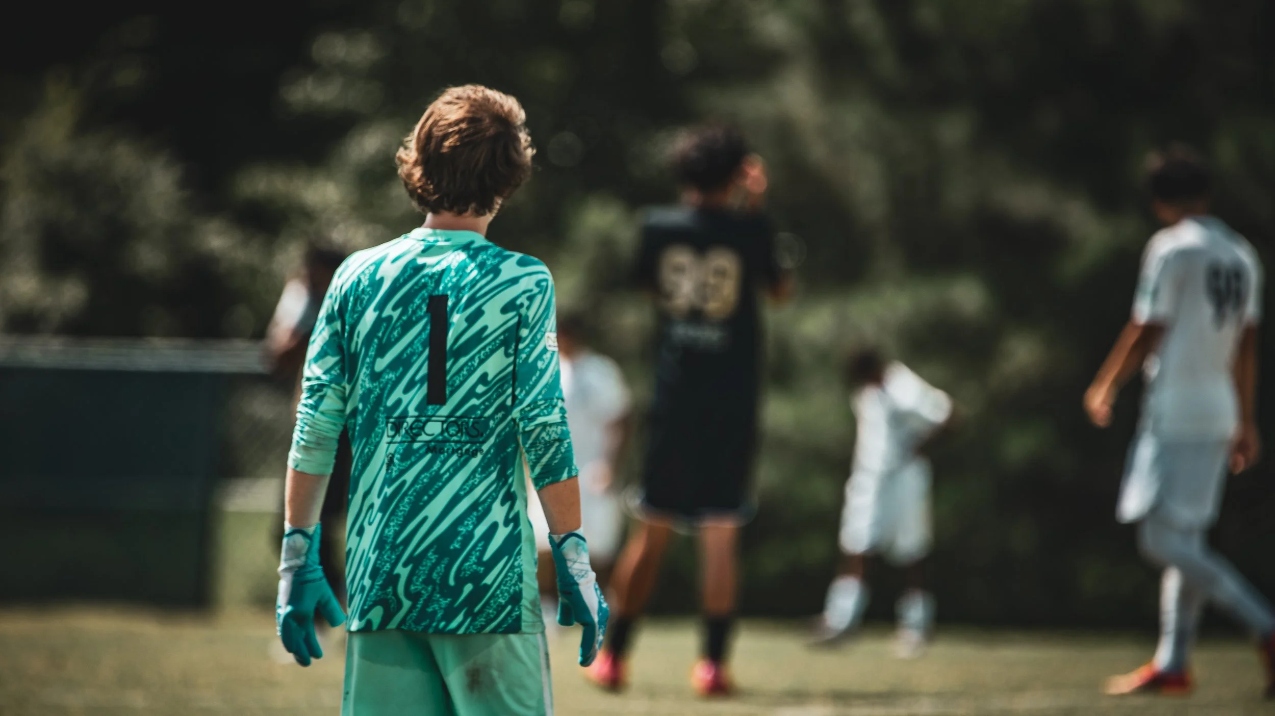 A soccer goalkeeper wearing a turquoise and black jersey with the number 1, standing on a soccer field, with other players and spectators in the background.
