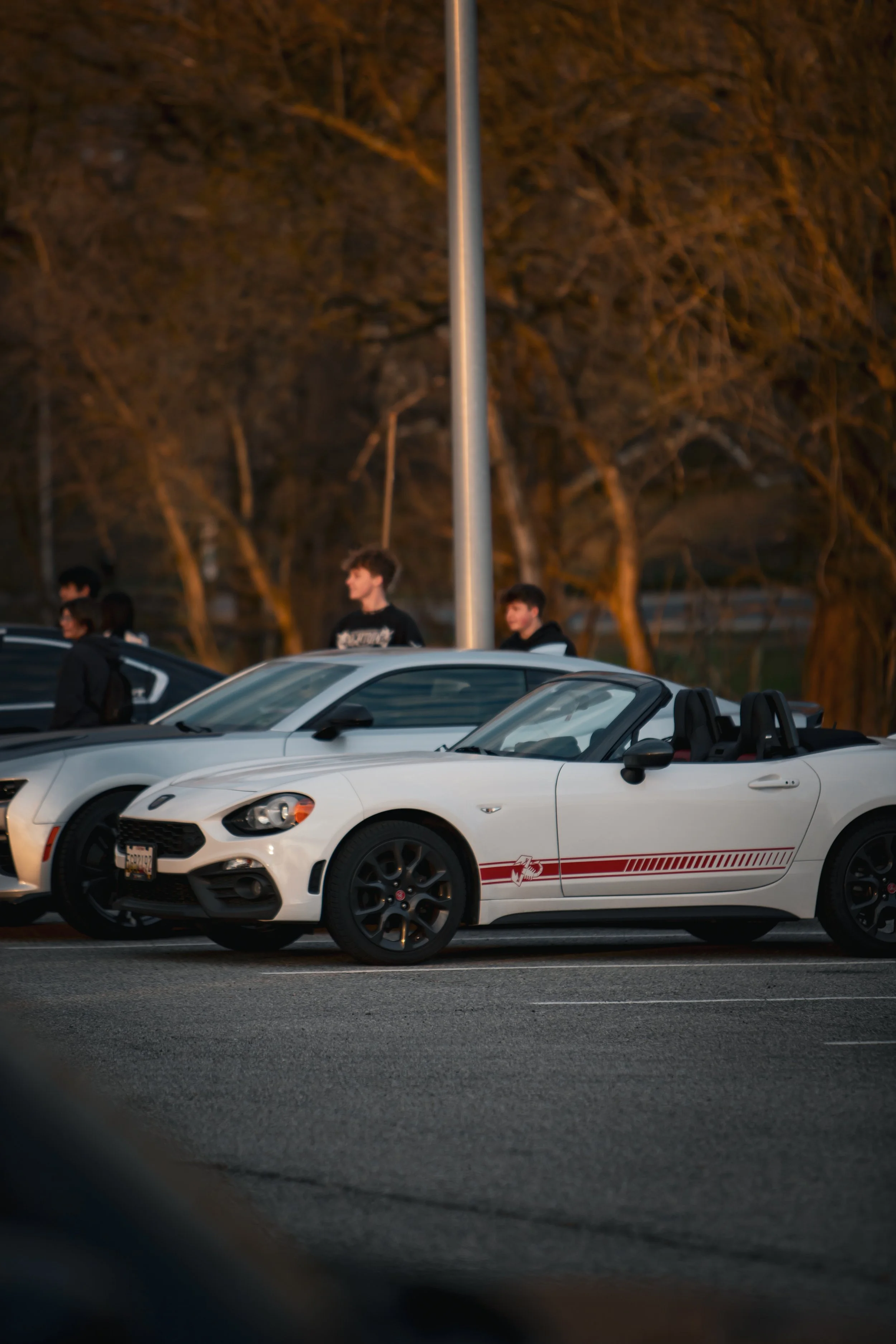 A parking lot at sunset with a white convertible sports car in the foreground and several people standing near other vehicles in the background.