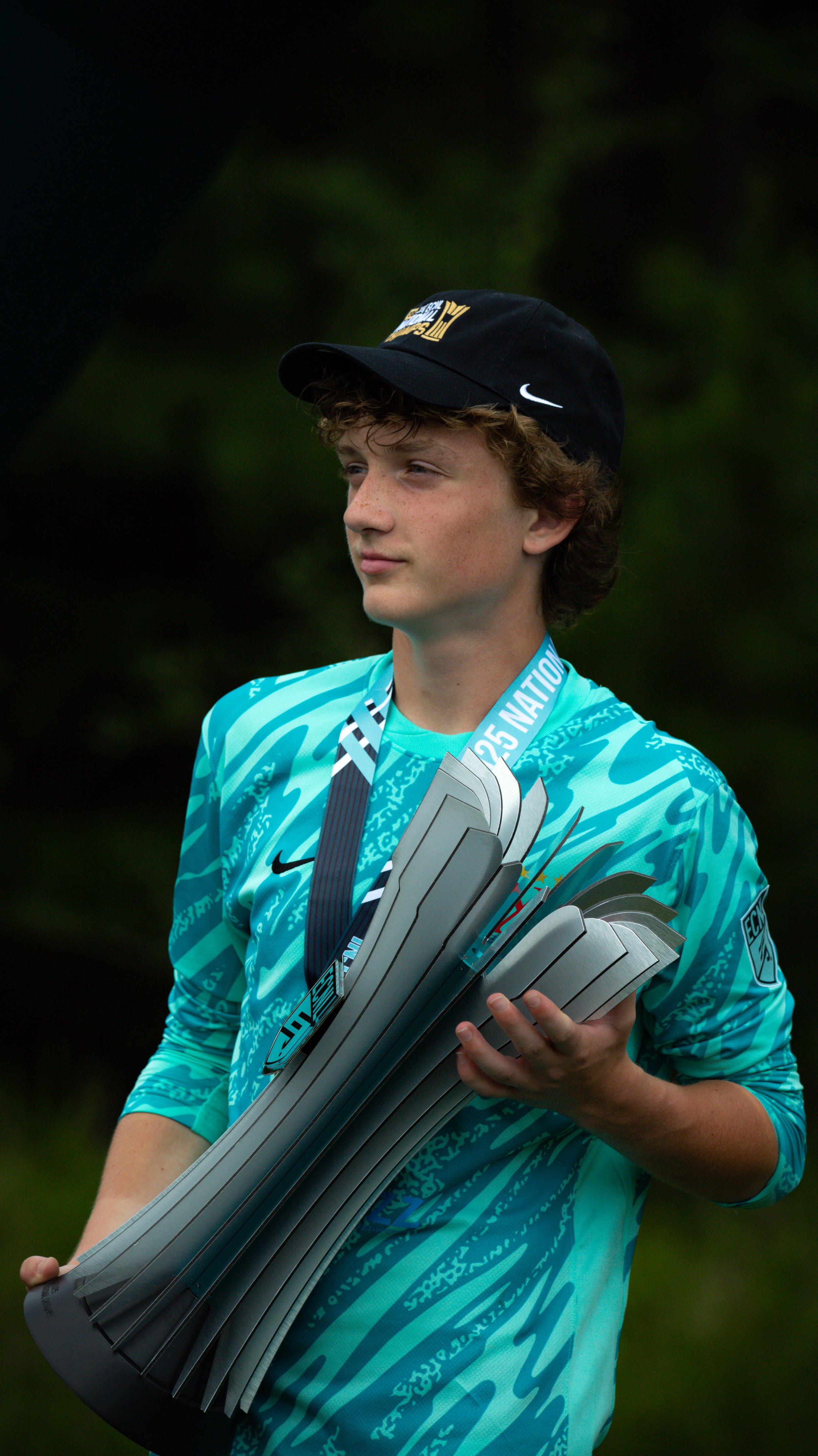 A young male athlete holding a trophy and medals, wearing a teal sports jersey and a black cap, standing outdoors with a blurred green background.