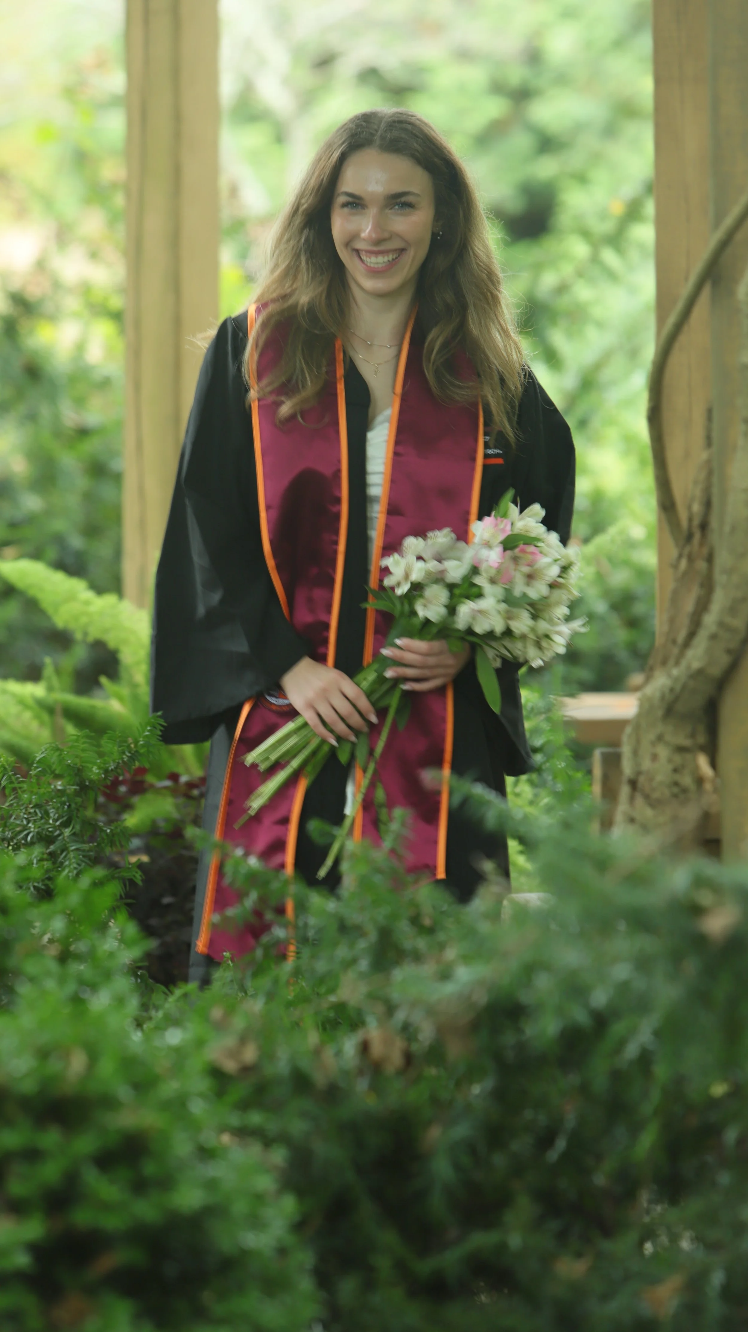 A woman in a graduation gown and cap holding a bouquet of flowers, smiling outdoors in a lush green setting.