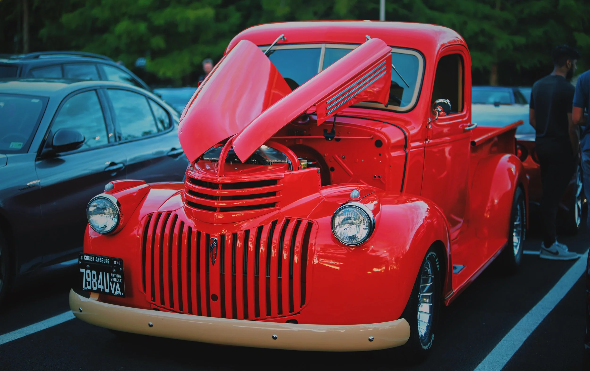 A vintage red pickup truck with its hood open, displayed at a car show, parked next to a black modern car.