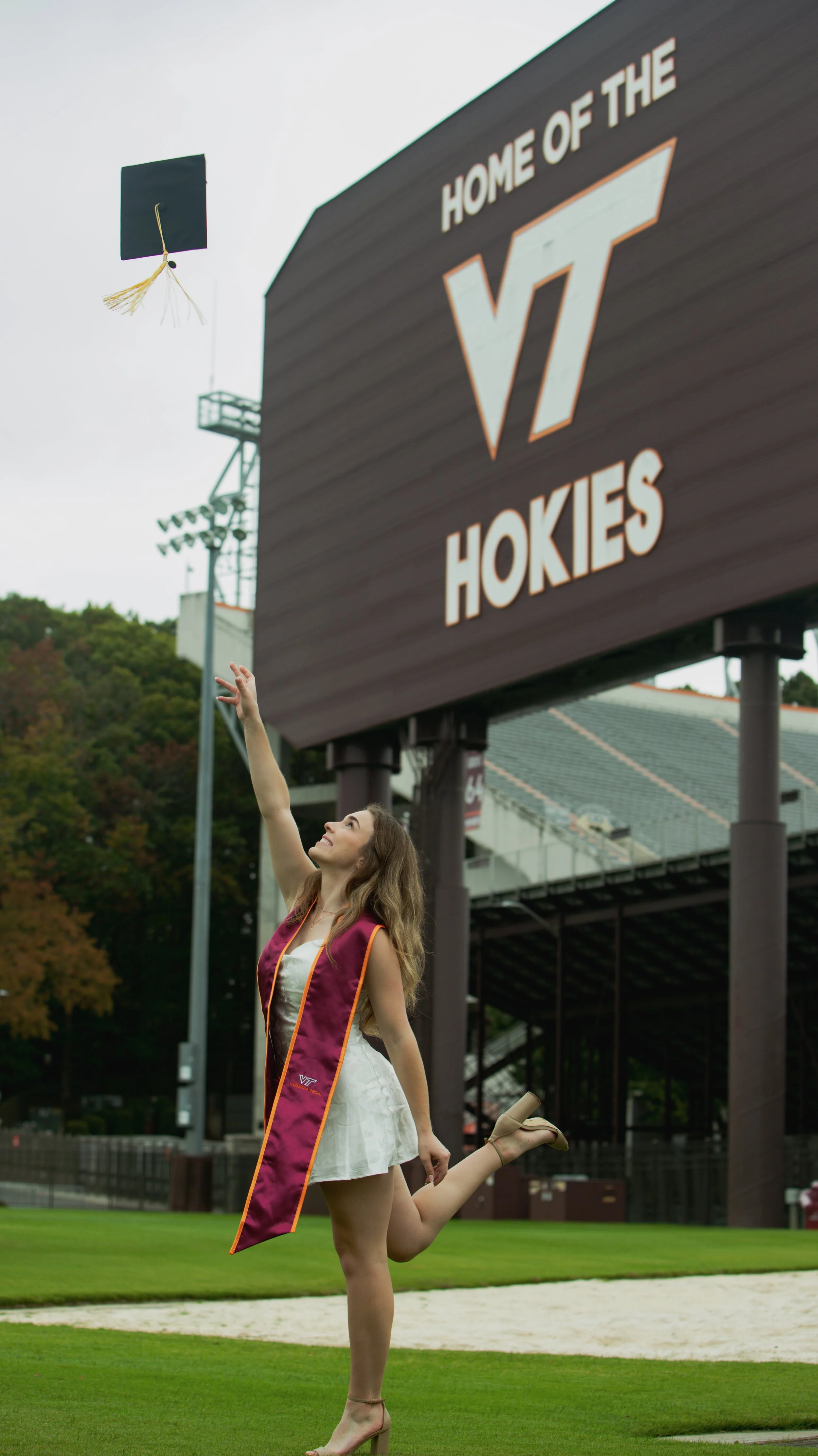 A young woman in a white dress and maroon graduation stole, celebrating on a football field after graduation, with a Virginia Tech Hokies sign and a graduation cap flying in the air.