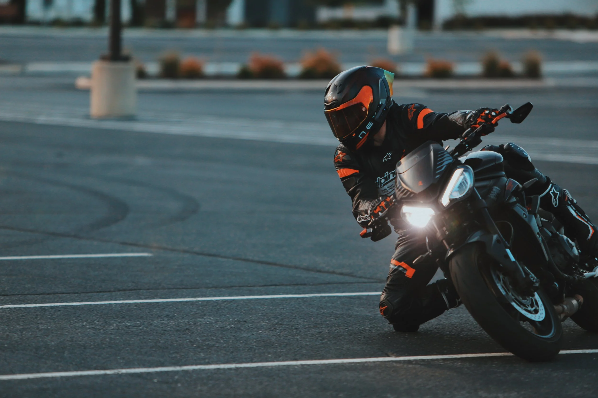 Motorcycle rider wearing black and orange gear leaning into a turn on an empty parking lot at dusk.