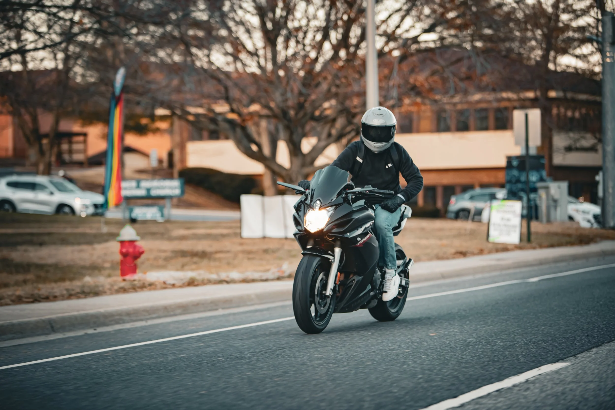 A motorcyclist wearing a white helmet and black jacket riding a black motorcycle on a city street with trees and buildings in the background.
