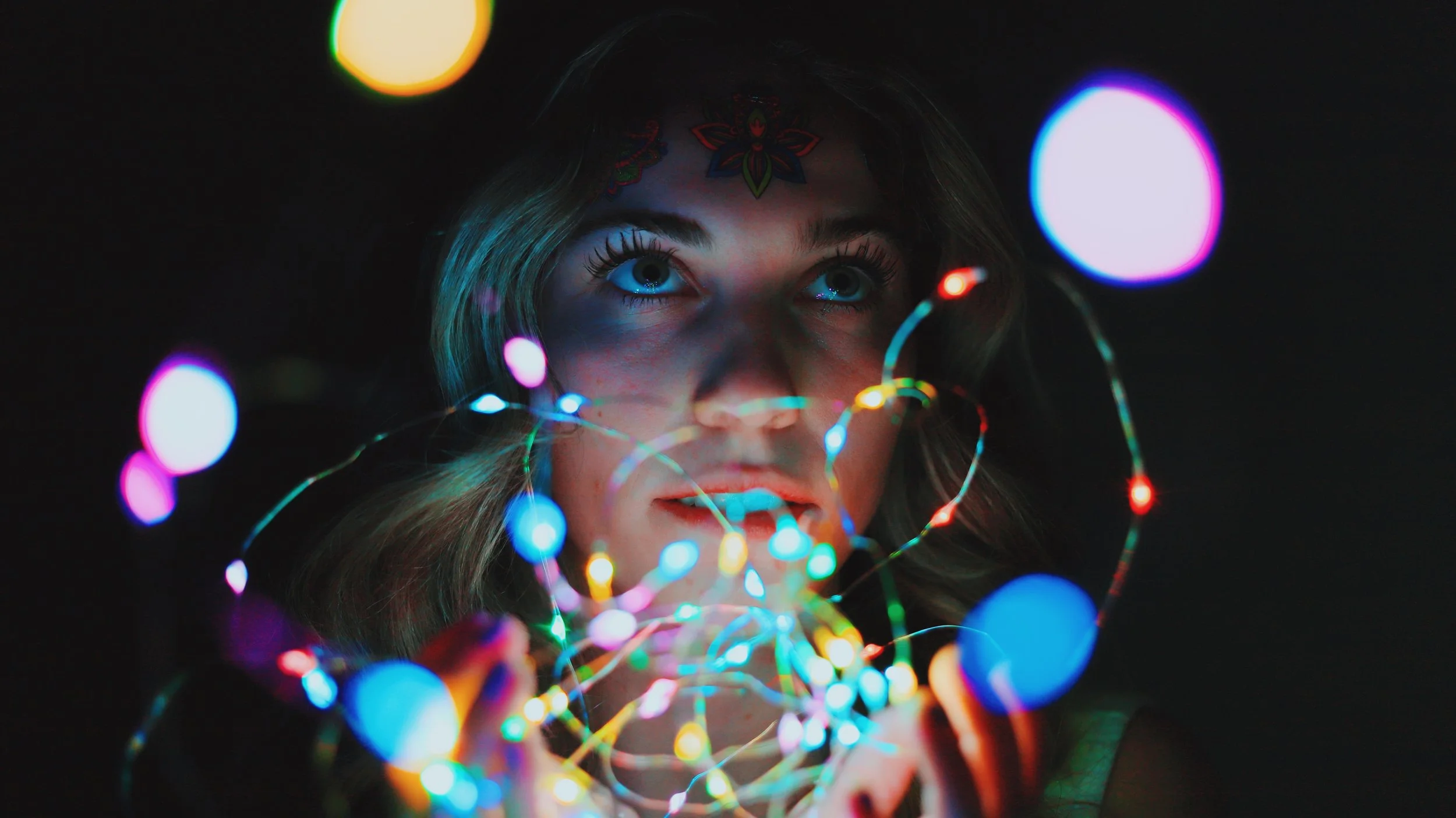 Young woman with colorful LED lights in her hands, looking up with illuminated face, dark background.